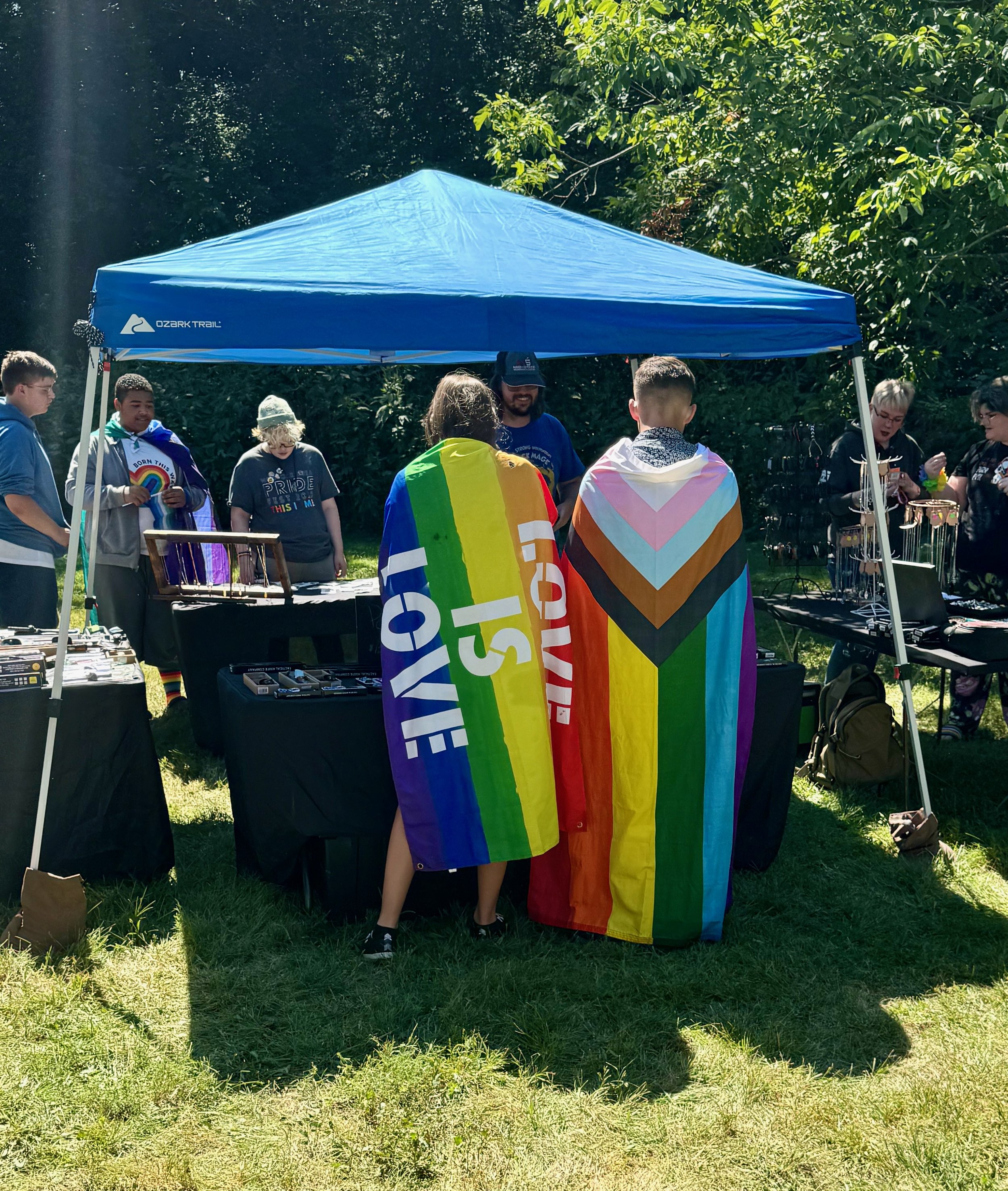 People at vendor booth with Pride flags draped as capes. 