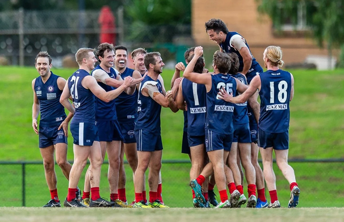 Group of male Australian rules football players celebrating on the field, wearing blue jerseys and shorts, with some lifting a teammate in the air.