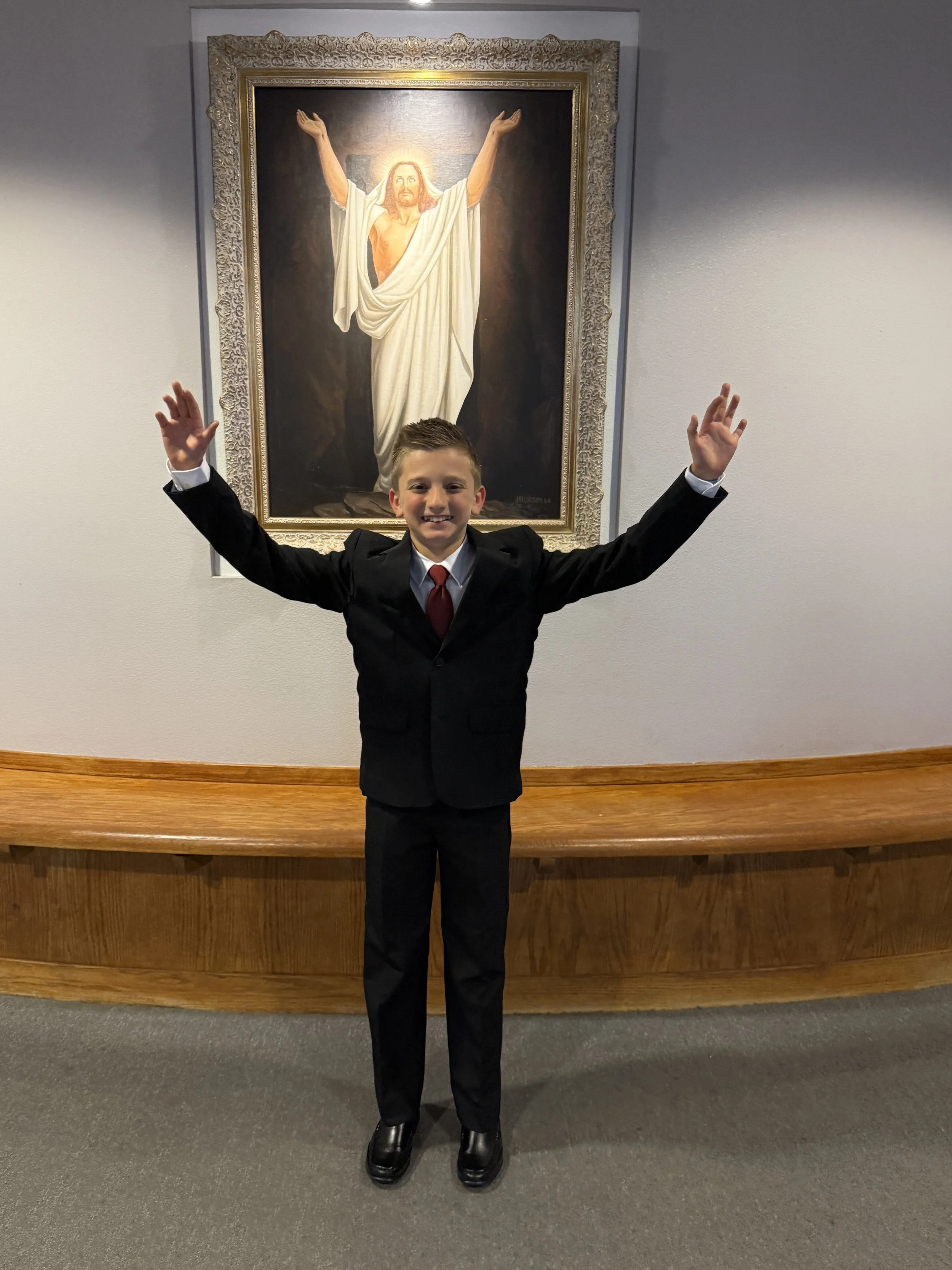 A young boy dressed in a black suit with a red tie standing in front of a painting of Jesus Christ with arms raised, inside a room with white walls and wooden trim.