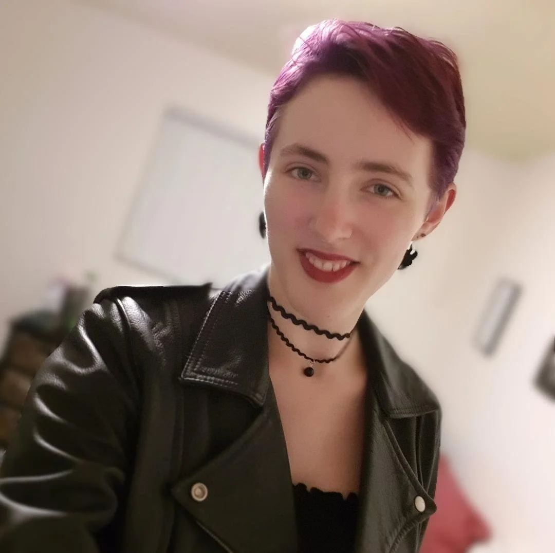 A young woman with short purple hair taking a selfie indoors, wearing a black leather jacket, black choker necklaces, and black earrings, smiling at the camera.