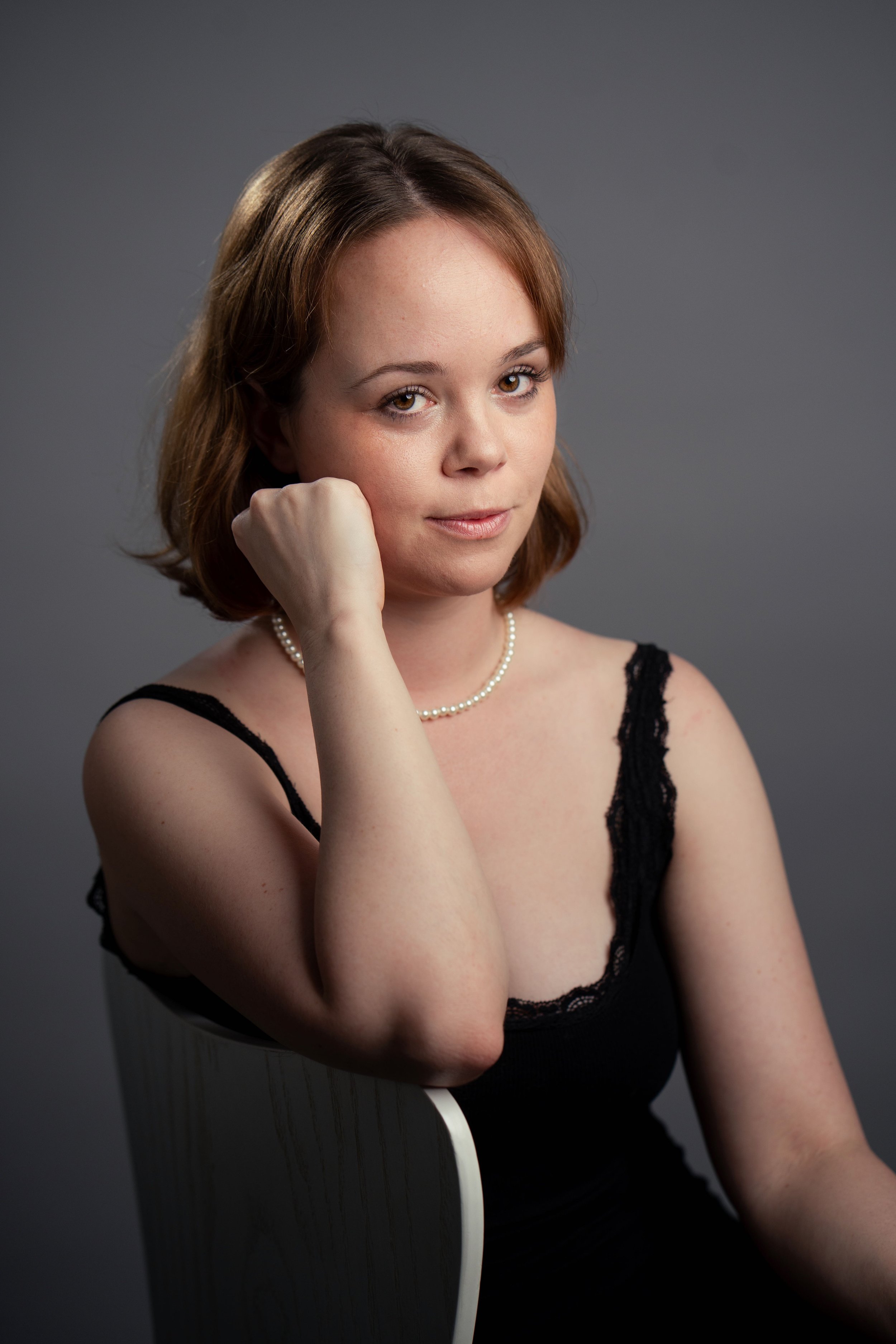 Portrait of a woman with shoulder-length brown hair wearing a black sleeveless top and pearl necklace, sitting against a gray background.