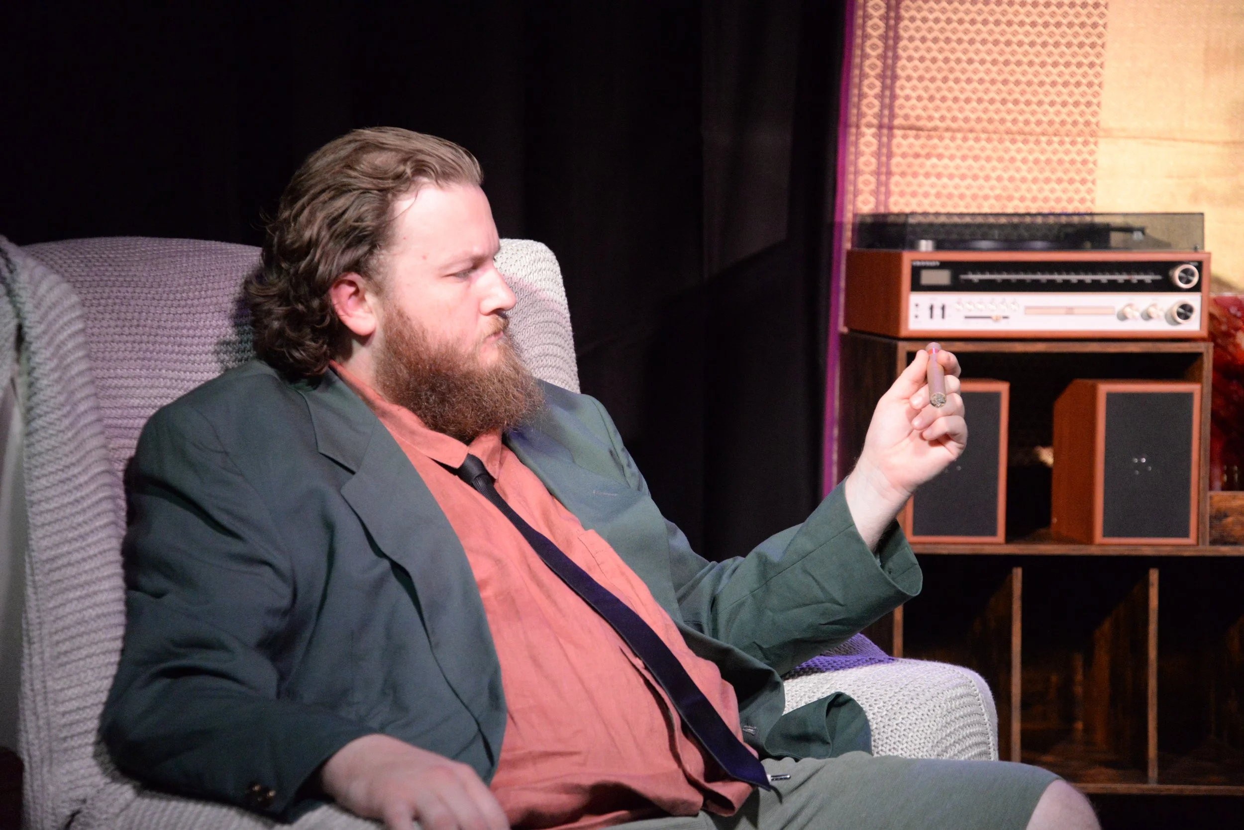 A man with long hair and a beard sitting on a couch, holding a lit cigar, and looking to the side, with vintage stereo equipment in the background.