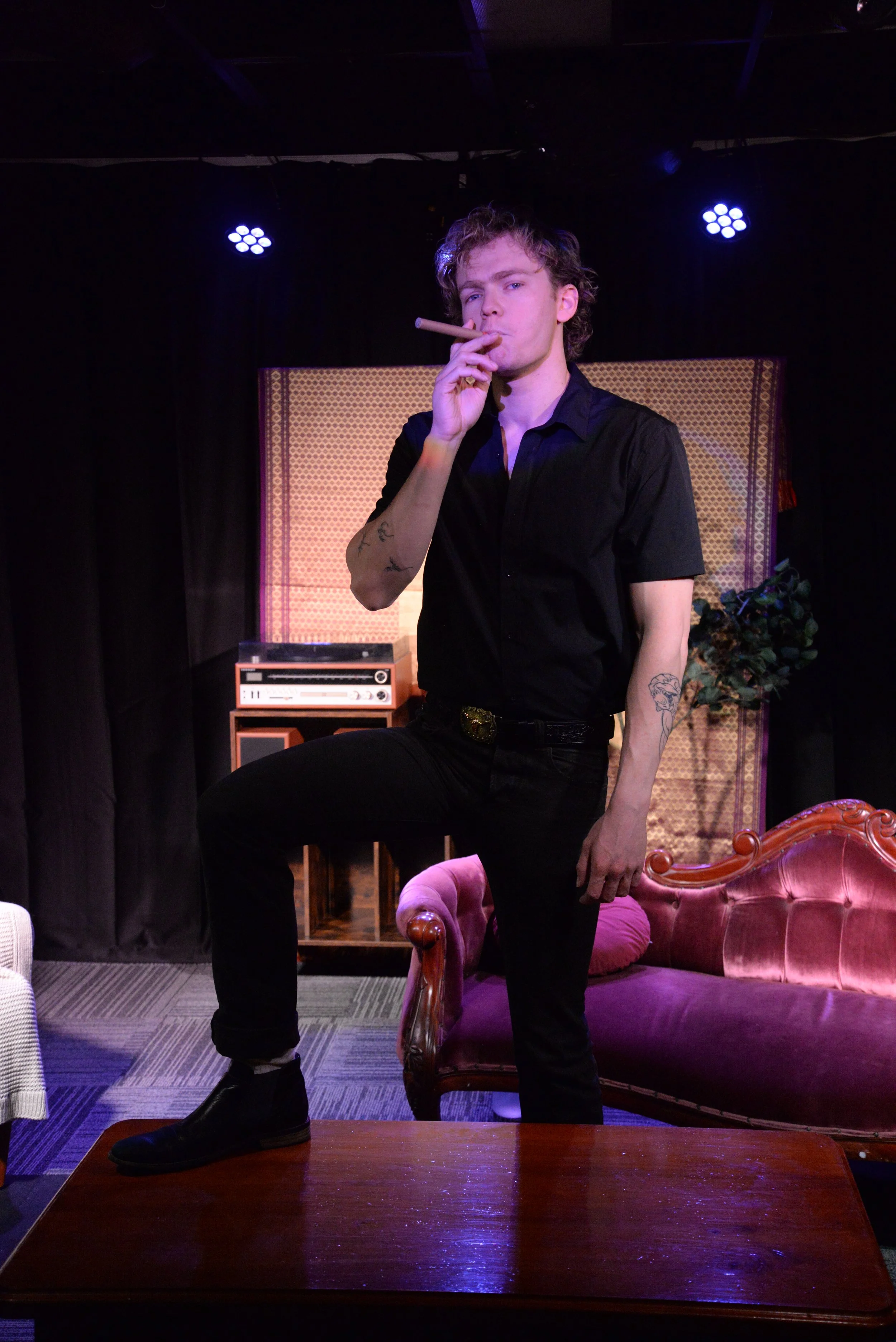 A young man with curly hair stands with one foot on a pink velvet armchair, smoking a cigar, in a room with vintage decor, including a pink velvet sofa, an old stereo, and a backdrop with a patterned design.