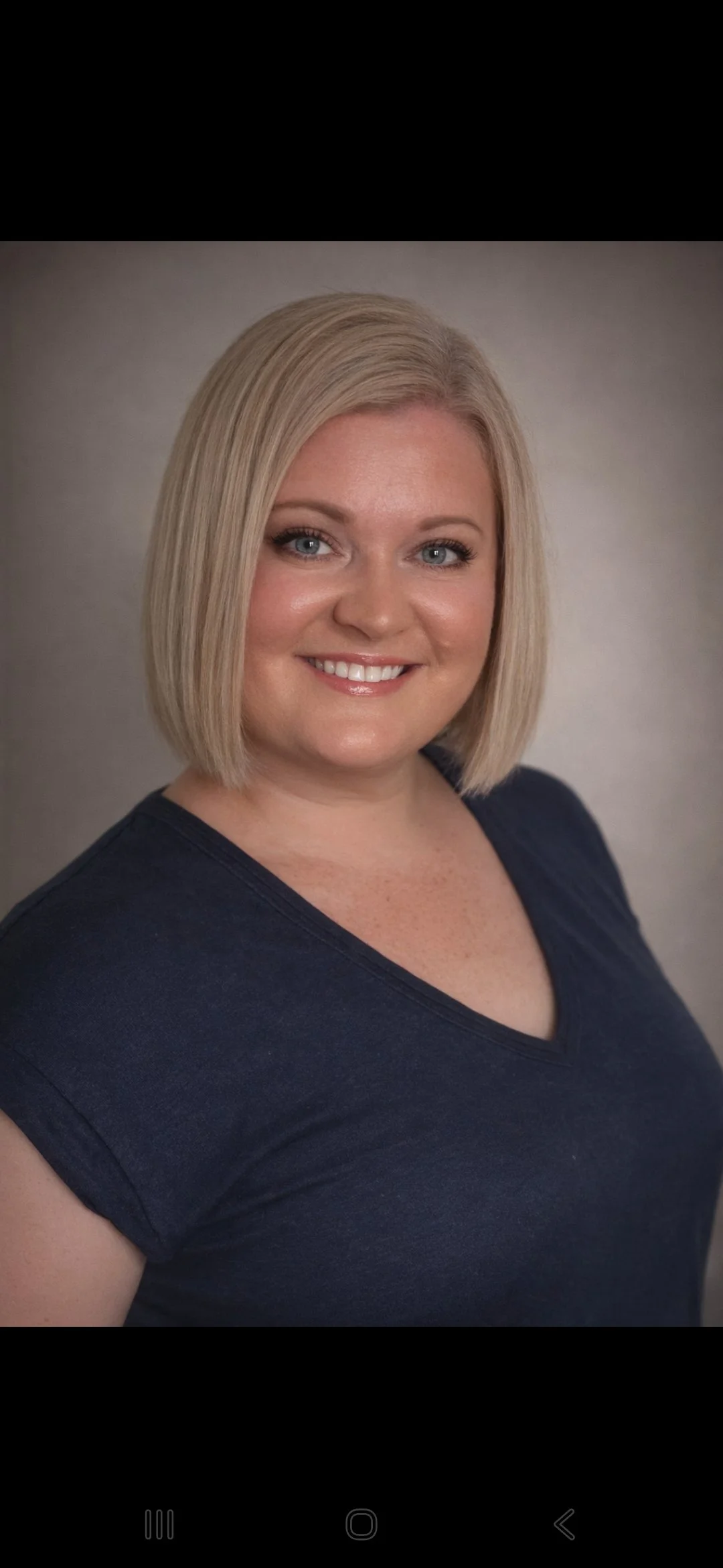 A smiling woman with shoulder-length blonde hair, wearing a navy blue shirt, posing against a neutral background.