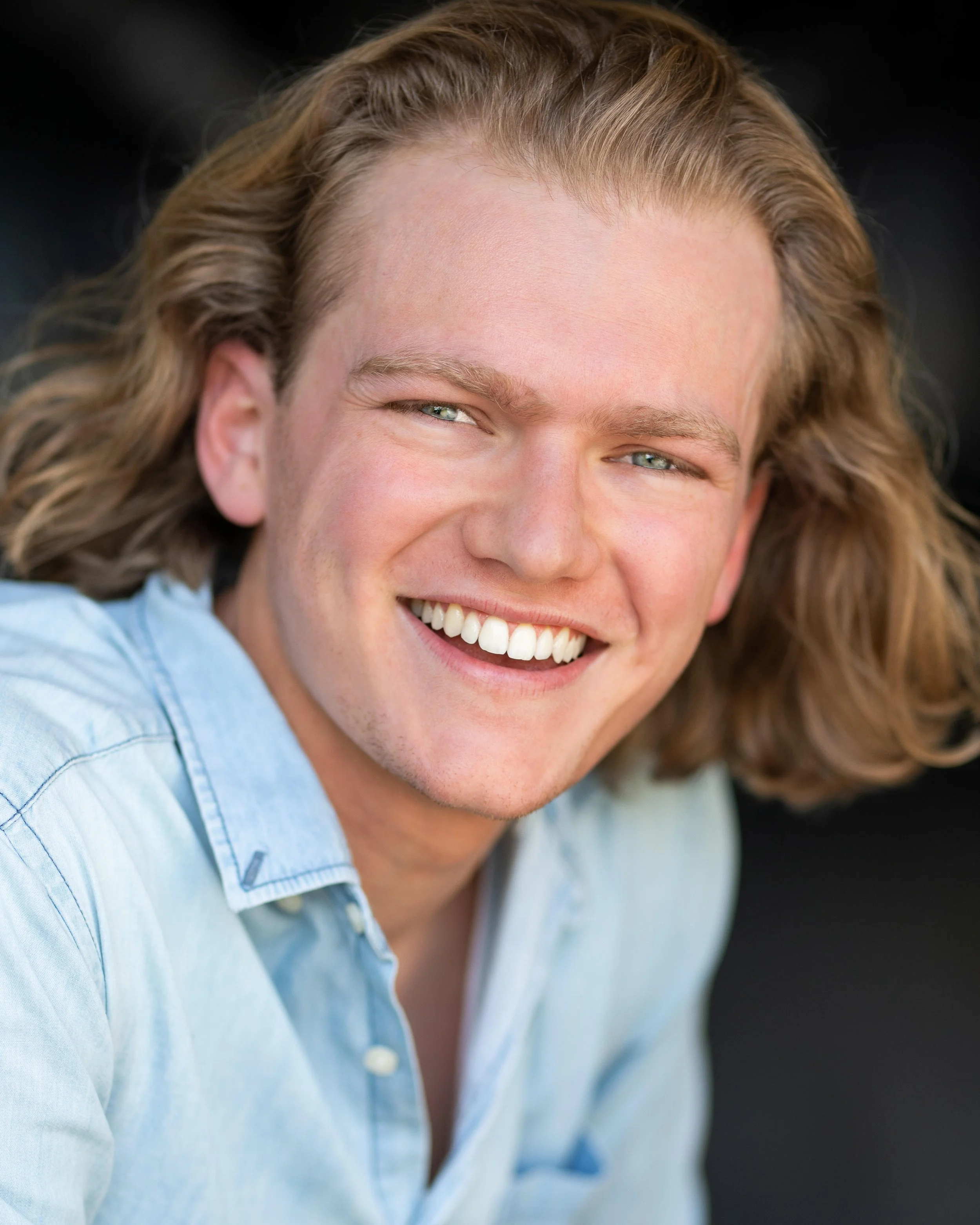 A young man with long blond hair, smiling broadly, wearing a light blue shirt.