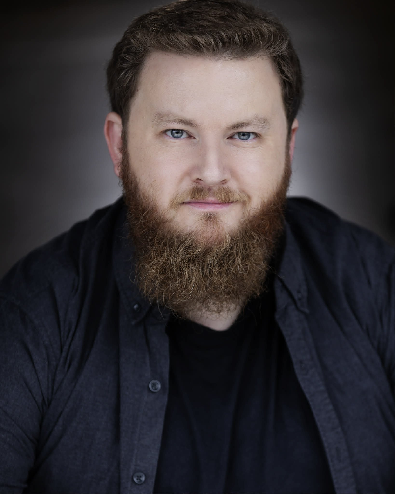 A headshot of a man with light skin, blue eyes, and a full beard, wearing a dark shirt against a dark background.