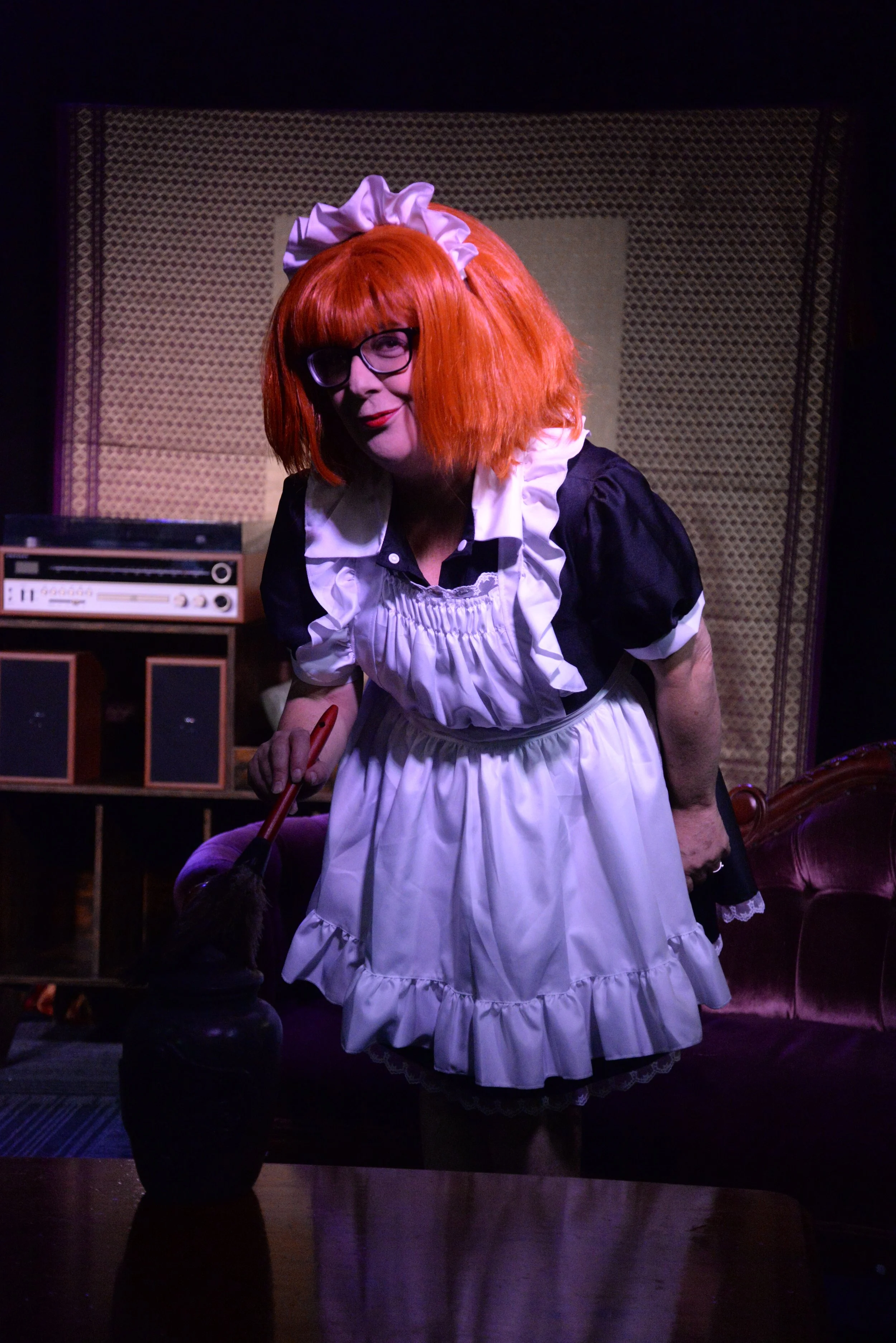 Person dressed as a maid with red hair, glasses, and black and white costume, leaning over a table with a broom in a dimly lit room with vintage furniture and a stereo in the background.