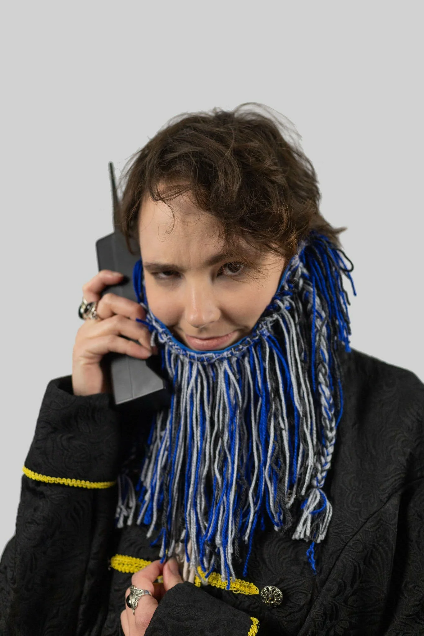 Person with curly hair holding a phone to their ear, wearing a black jacket with yellow details and a fringed blue, black, and white scarf.