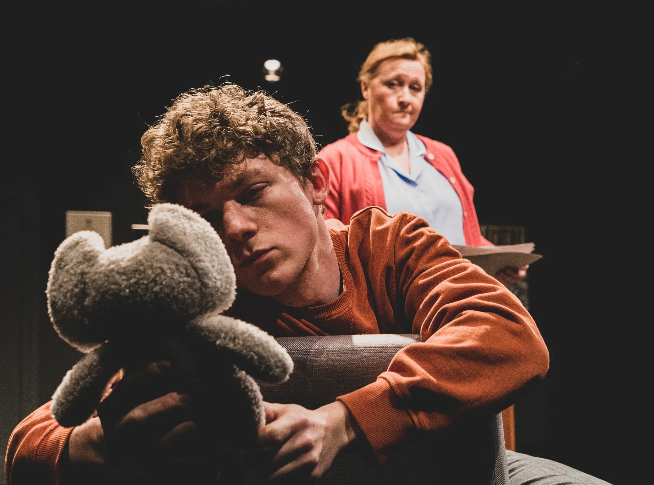 A young man with curly hair resting his head on a teddy bear, sitting on a chair, with an older woman standing behind him holding a clipboard, in a dark room.