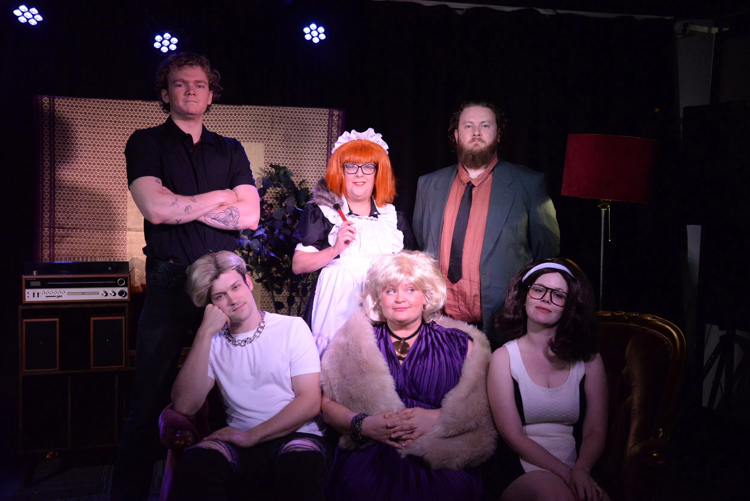 Group of six people in costumes posing for a photo on stage, some sitting and some standing, with stage lighting overhead and a backdrop behind them.