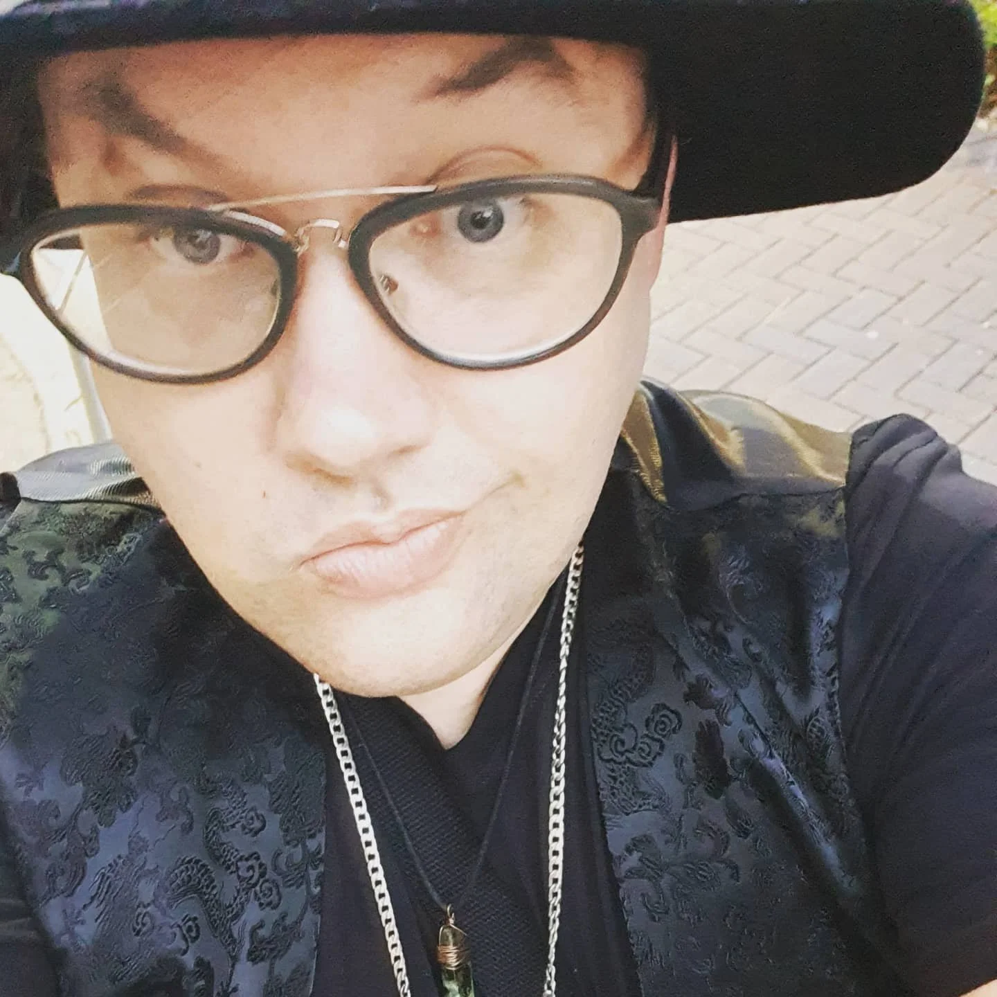 Close-up of a person wearing black-rimmed glasses, a black hat, and a patterned black jacket with gold accents, standing outdoors on a brick path.
