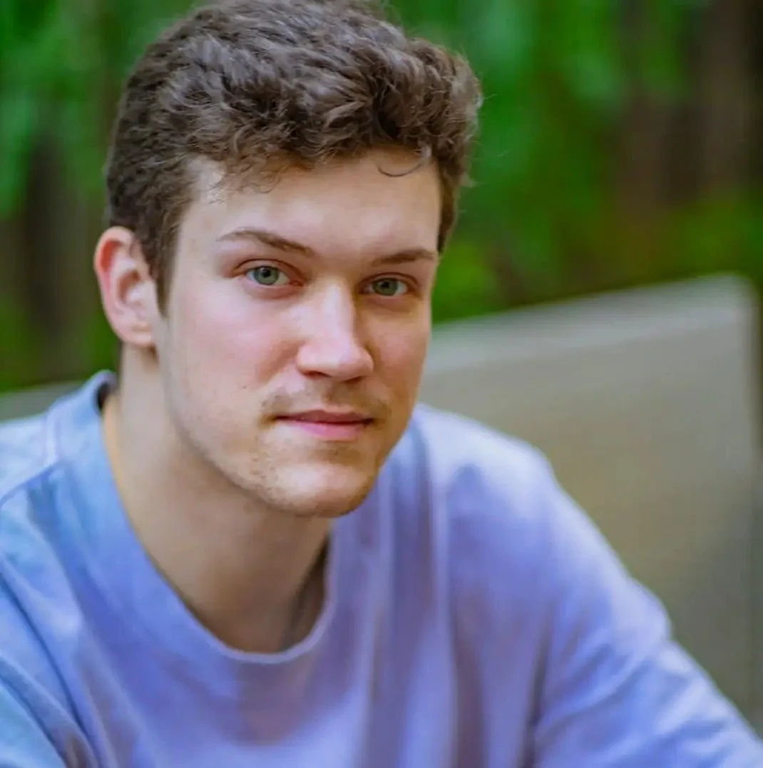 A young man with short curly brown hair and blue eyes, wearing a blue shirt, sitting outdoors with blurred green trees in the background.