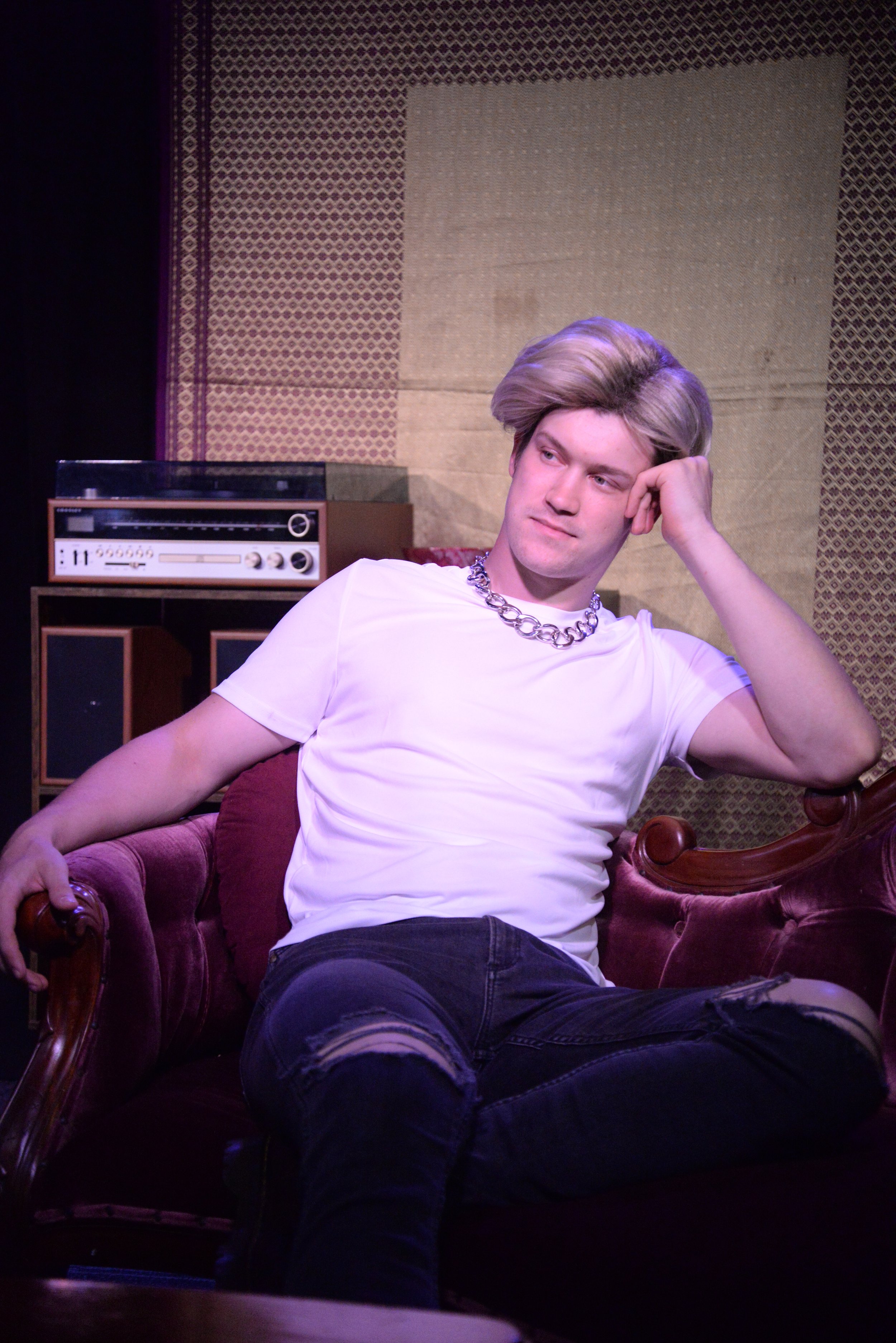 Young man with styled blond hair wearing a white T-shirt, ripped black jeans, and a chunky chain necklace, sitting on a vintage purple velvet sofa in a room with patterned wallpaper and a stereo in the background.