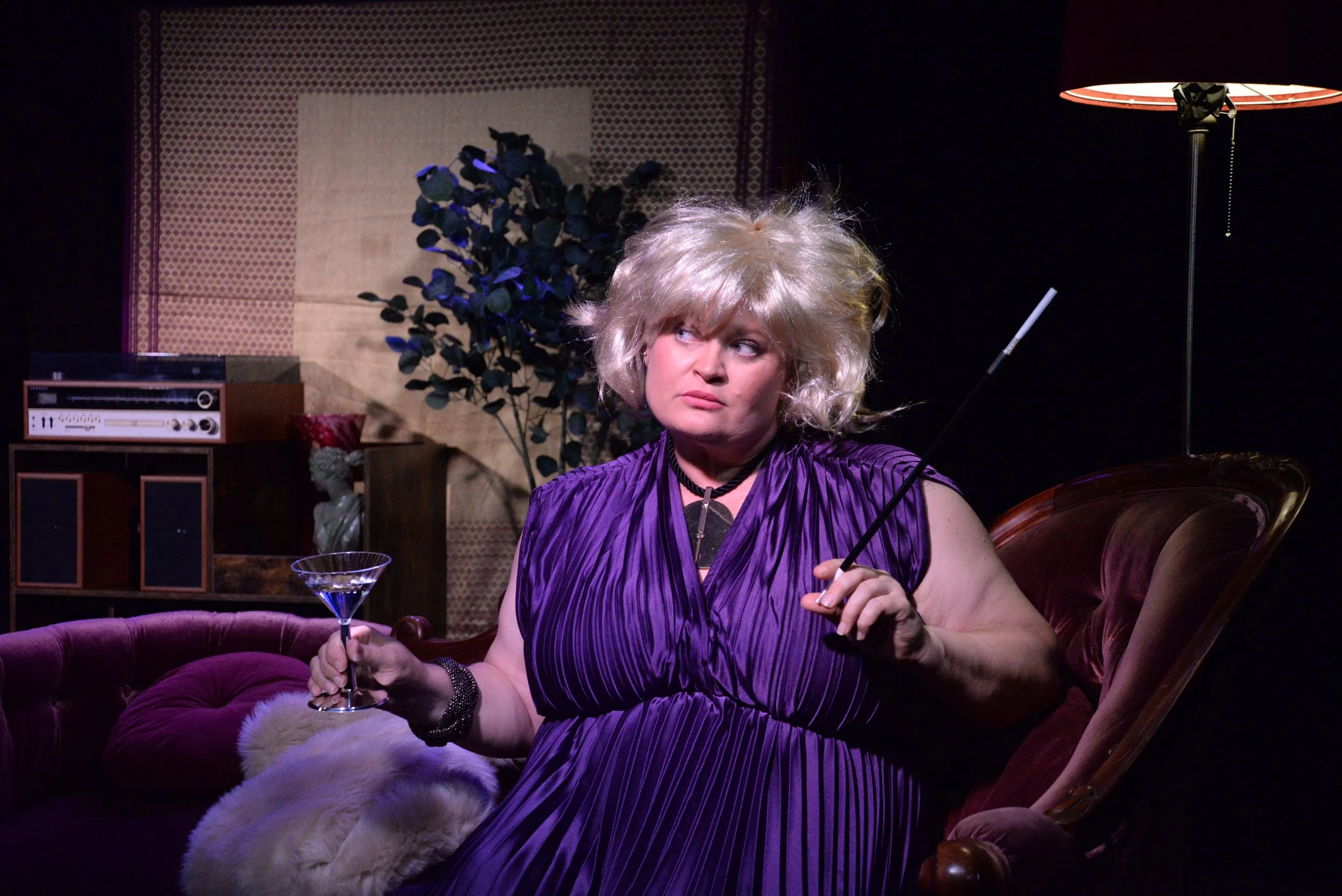 A woman with a blonde wig and purple dress sitting on a velvet couch, holding a martini glass and a cigarette holder, with a serious expression.