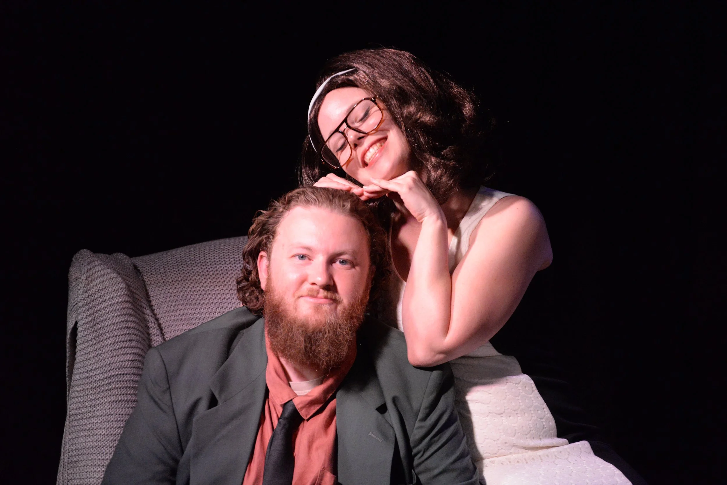 A man with a beard in a suit and a woman with glasses and black hair in a white dress, both smiling, sitting on a chair against a dark background.