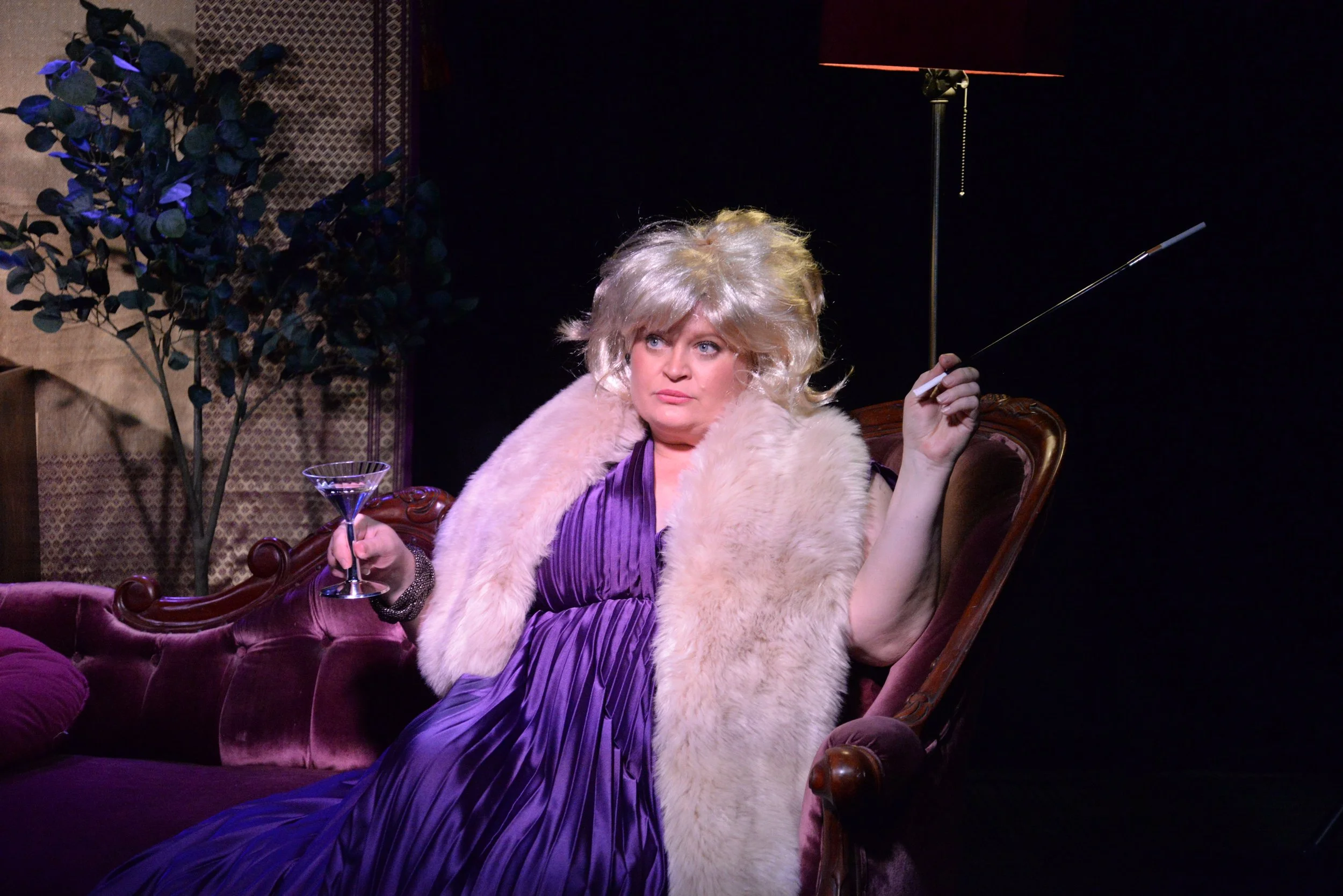 A woman sitting on a vintage sofa, dressed in a purple gown with a fur stole, holding a martini glass in one hand and an unlit cigarette in the other, in a dimly lit room.