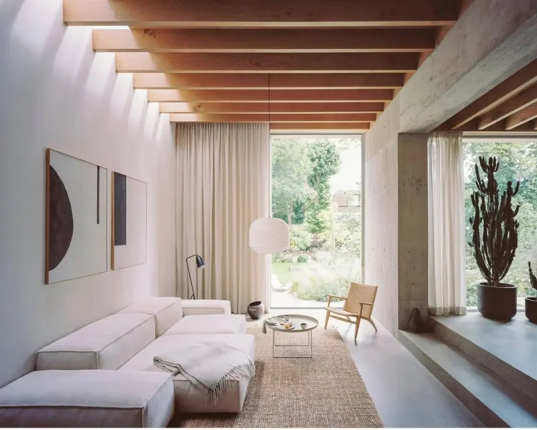 Modern living room with white sectional sofa, abstract wall art, wooden ceiling beams, large windows with curtains, wicker chair, potted cactus plant, and natural light.