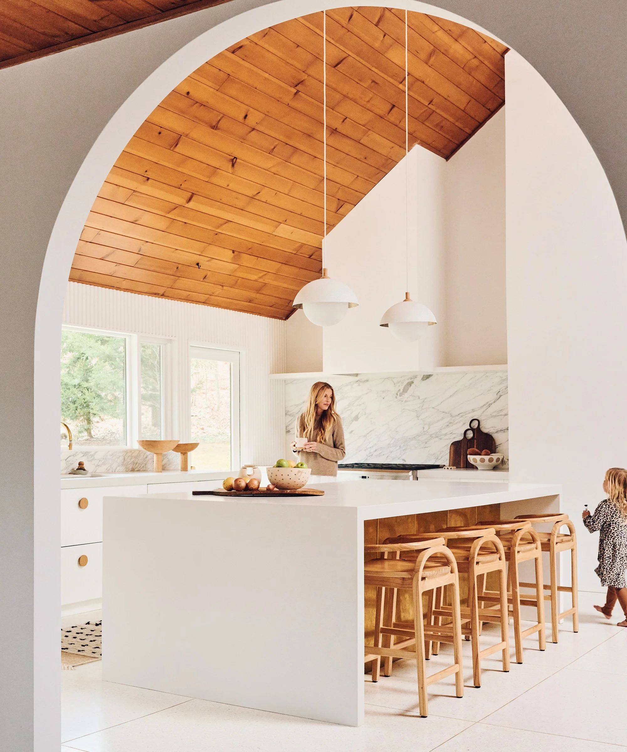 Modern kitchen with wooden ceiling, white cabinets, marble backsplash, and a white island with wooden barstools. A woman and a small child are present.