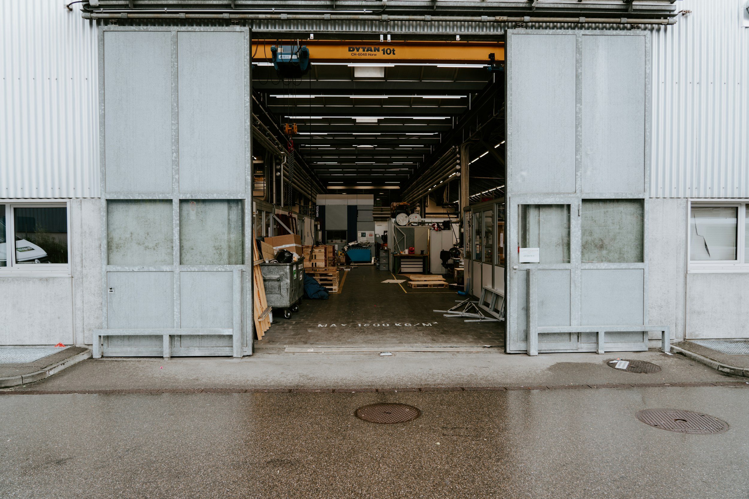View of a garage with open sliding doors, revealing an industrial workspace inside with various equipment, tools, and wooden pallets. The ground outside appears wet, indicating recent rain.