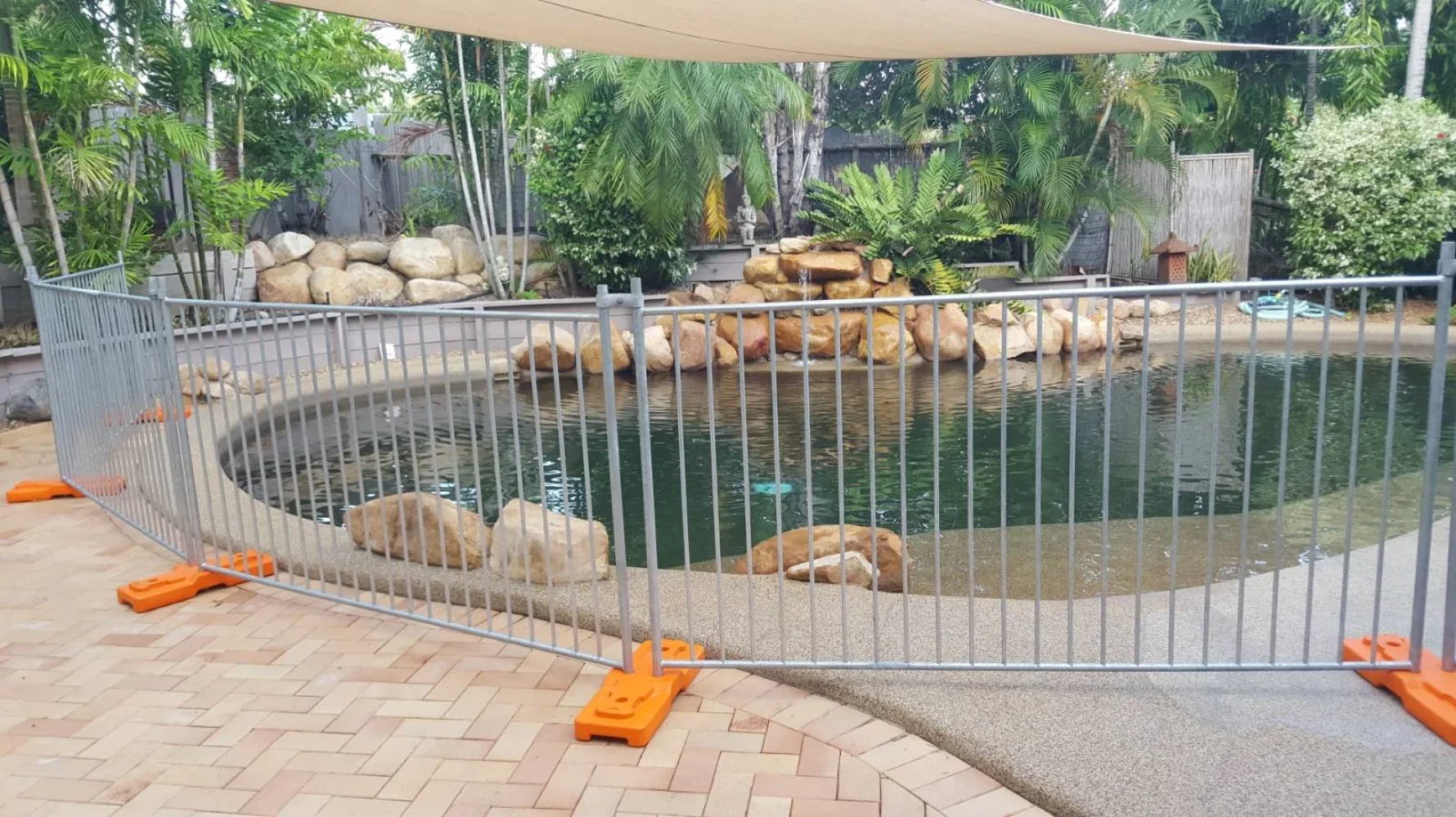 A backyard with a small pond enclosed by a metal safety fence, surrounded by lush green plants and trees, with rocks and a decorative waterfall feature.