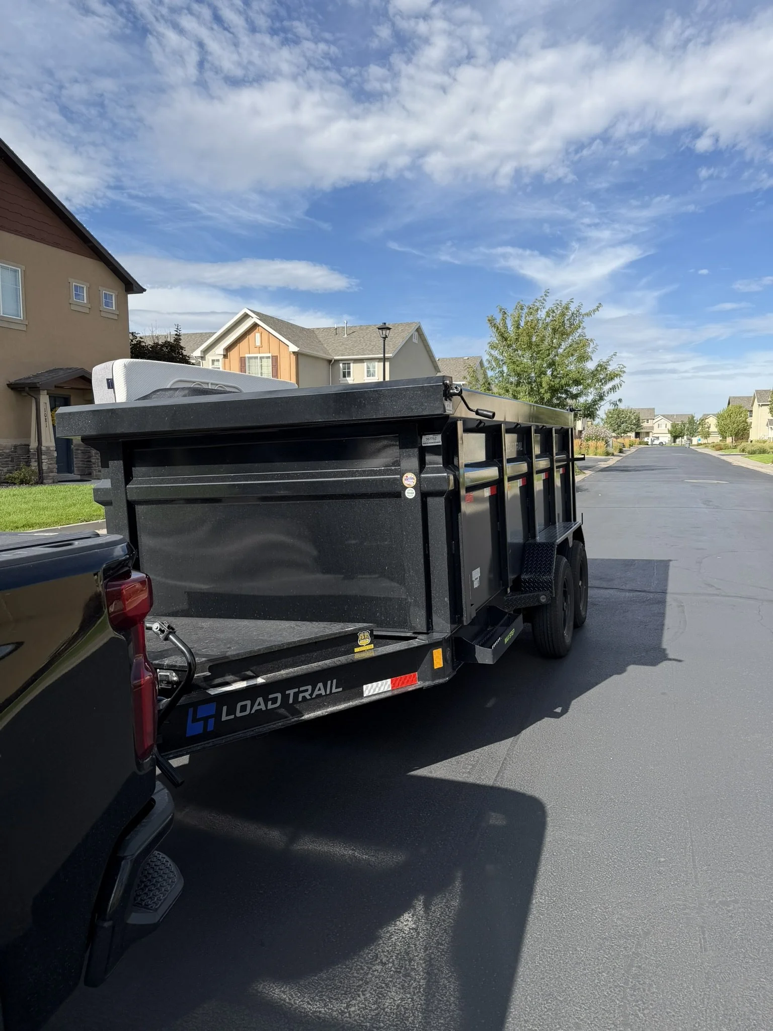 A black flatbed truck parked on a suburban street, with a mattress and some furniture on it. The street has houses with trees and a partly cloudy blue sky.