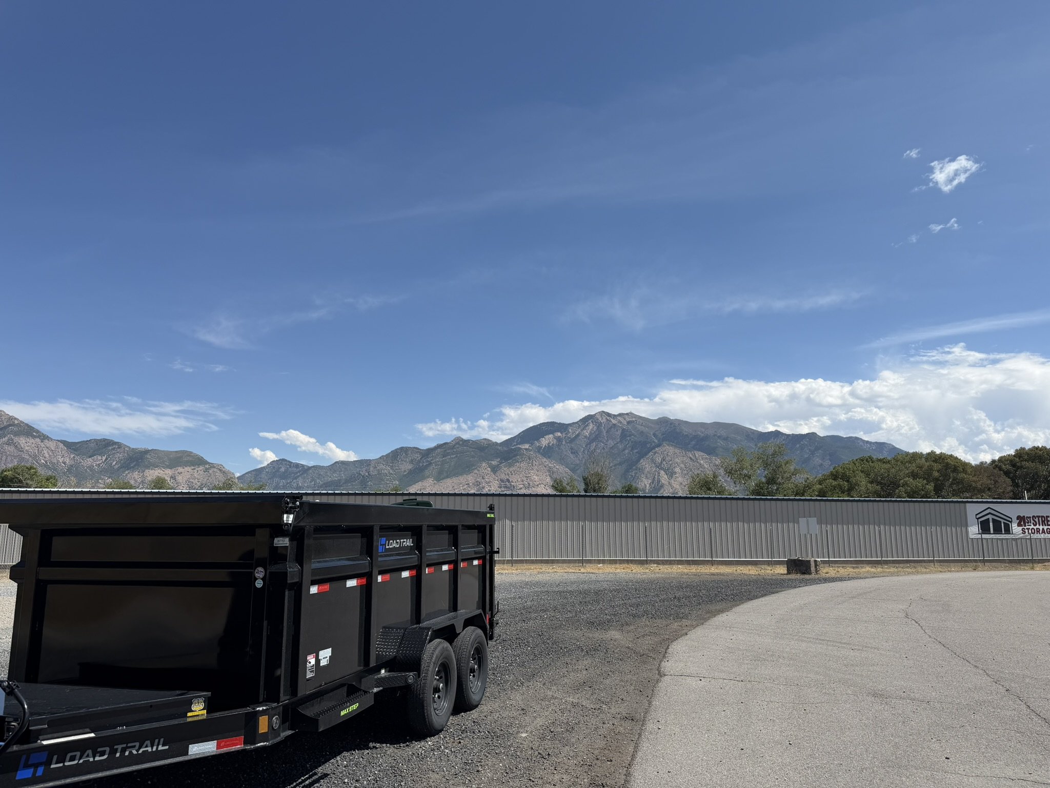 Black utility trailer parked outside a large gray building with a mountain range in the background under a blue sky with clouds.