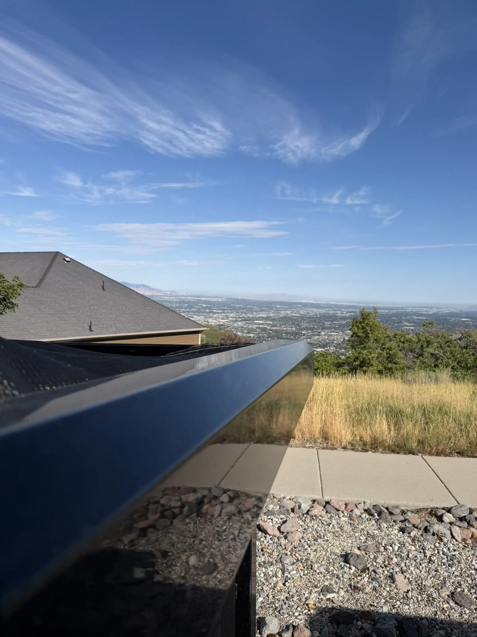 A view from a backyard showing a rooftop, distant cityscape, blue sky with wispy clouds, and a patch of grassy and rocky ground.