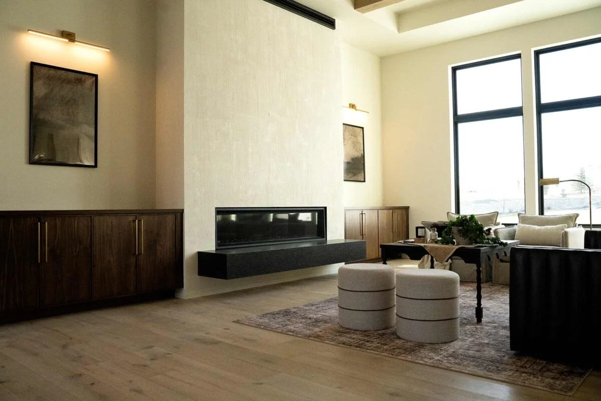 Modern living room with large windows, white walls, wooden flooring, a fireplace, and contemporary furniture including a sofa, armchair, and ottomans.