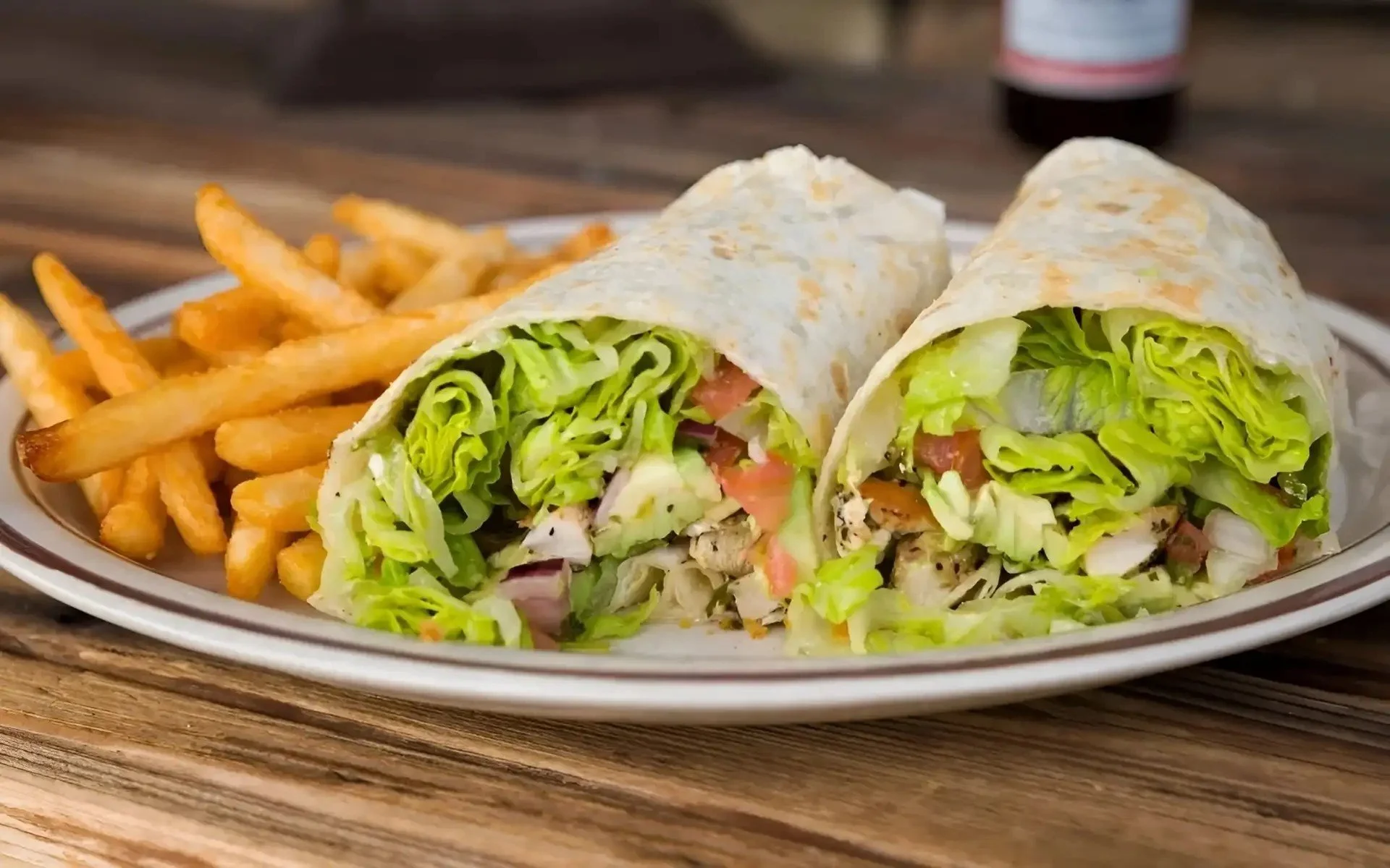 A plate of chicken Caesar wrap cut in half, with shredded lettuce, tomatoes, grilled chicken, and wrap, served with French fries.