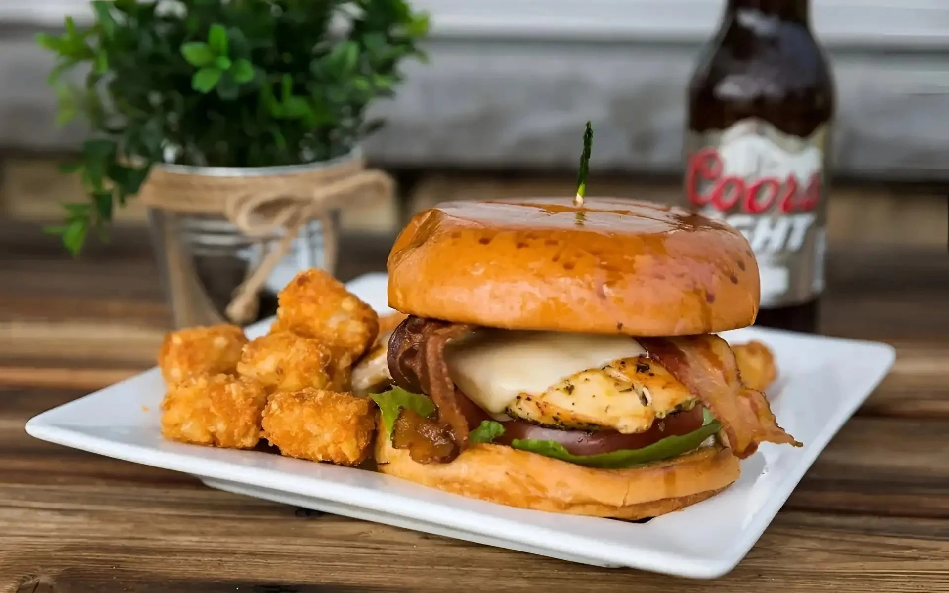 Sandwich with chicken, bacon, peppers, and cheese on a bun, served with fried chicken bites and a beer bottle in the background.
