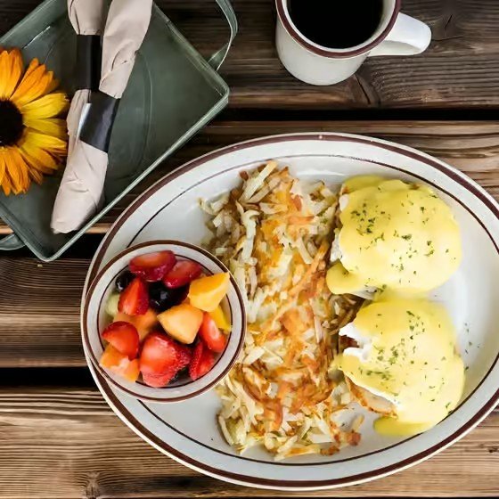 A breakfast plate with eggs Benedict, hash browns, and a side bowl of mixed fruit, along with a cup of coffee on a wooden table.