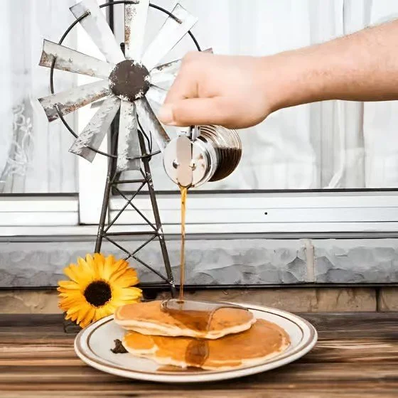 A plate with pancakes topped with syrup, with a sunflower beside it, on a wooden table. In the background, there's a small metal windmill and a glass of syrup being poured from a spoon.
