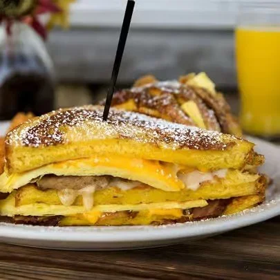 A plate with a breakfast sandwich made of eggs, cheese, and bacon between two crispy hash browns and topped with powdered sugar, served with a glass of orange juice in the background.
