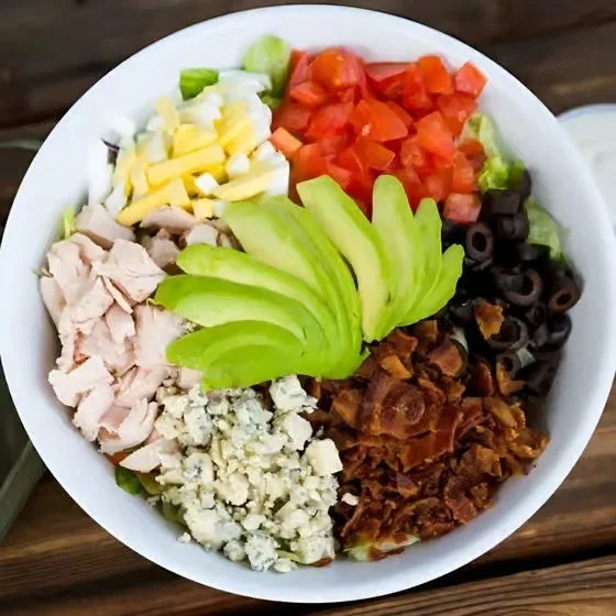 Bowl of salad with diced tomatoes, black olives, sliced avocado, chopped chicken, shredded cheese, bacon bits, and blue cheese crumbles.