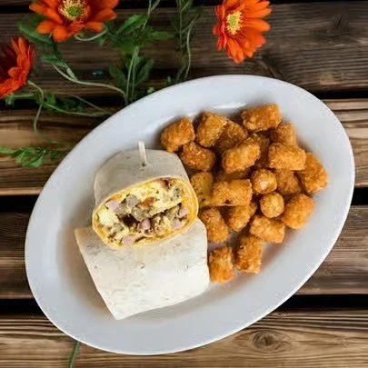 A white oval plate holding a breakfast burrito and a serving of tater tots on a wooden table with orange flowers nearby.