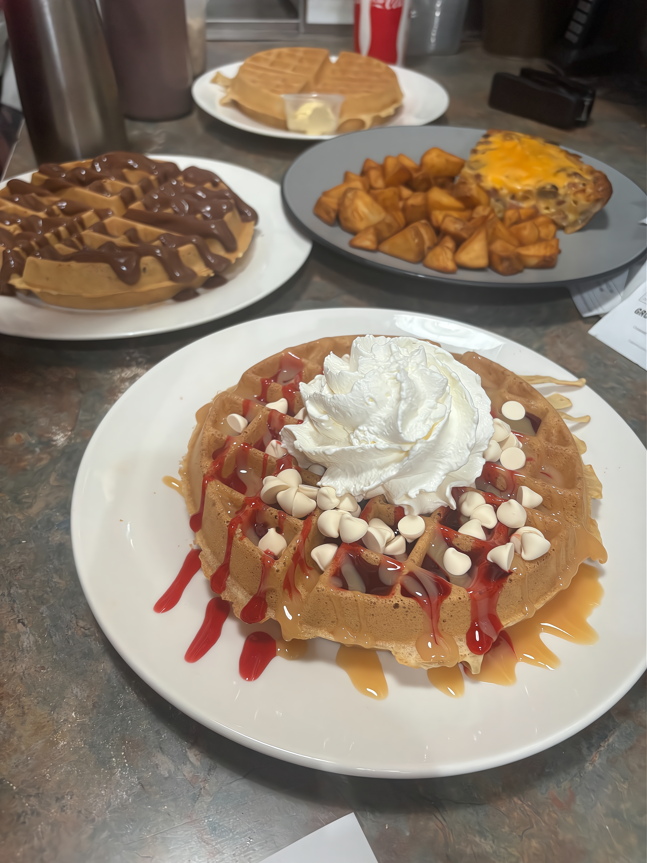 A plate of cinnamon roll topped with whipped cream, white chocolate chips, drizzle of caramel and red syrup, with additional food like waffles with chocolate syrup, potato wedges, and a slice of pizza in the background.