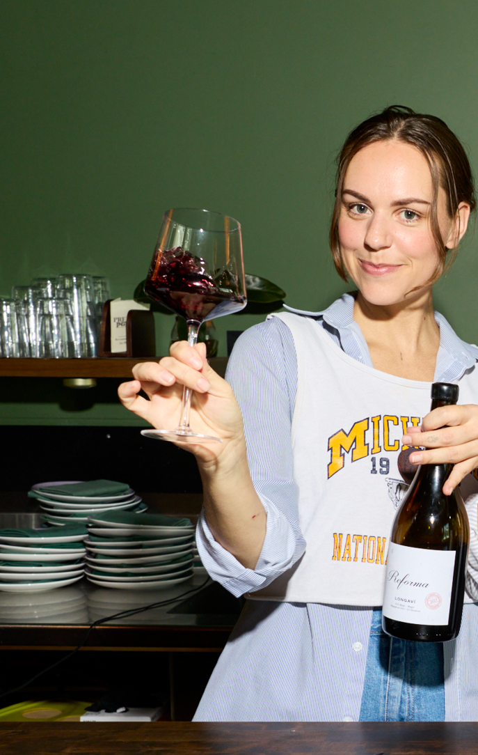A woman with short brown hair and fair skin holding a glass of red wine and a wine bottle, standing in a bar or restaurant setting with green walls and stacked dishes in the background.