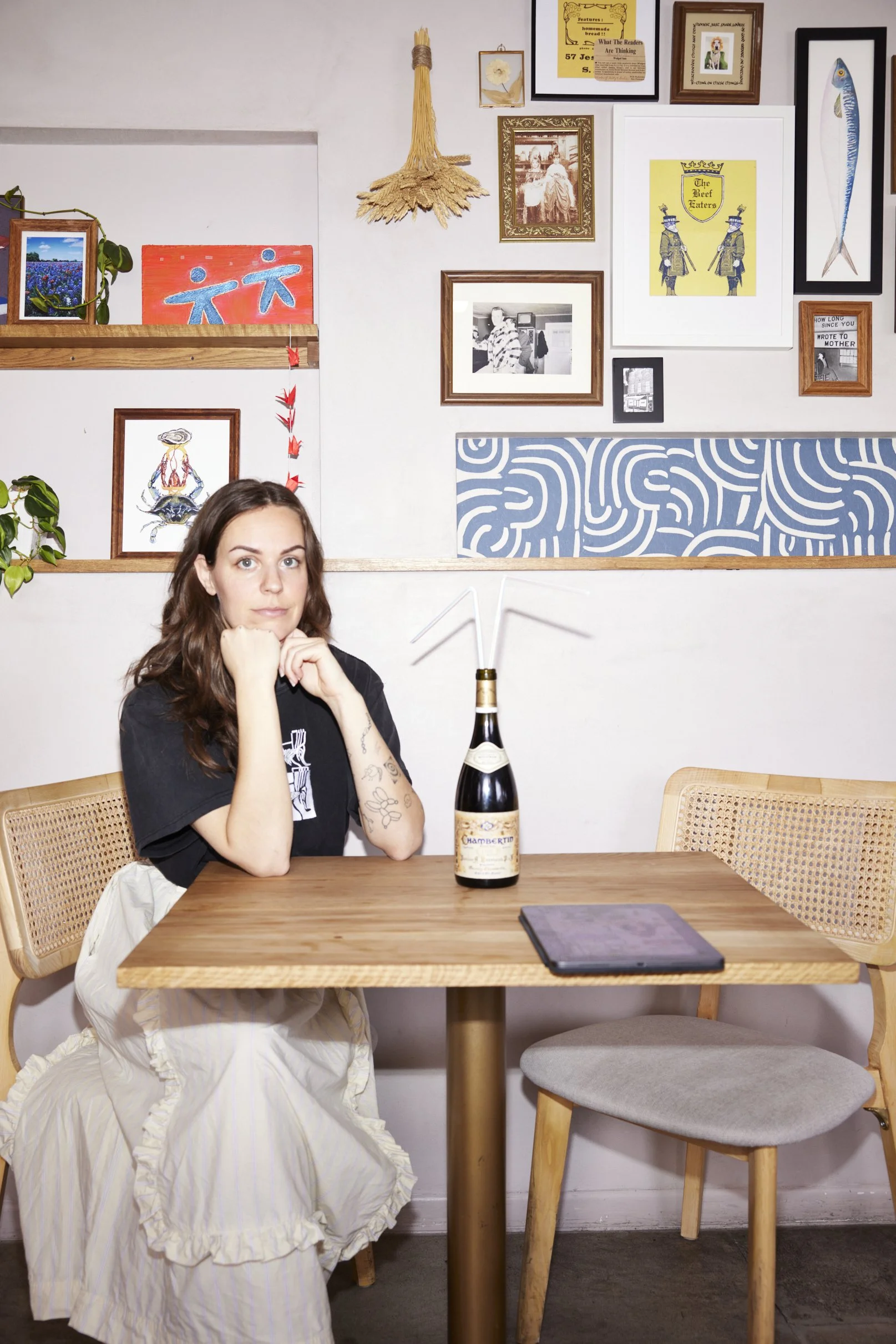 A woman sitting at a wooden table with a bottle of champagne and a tablet, in a room decorated with various framed pictures, artwork, and decor on the wall.