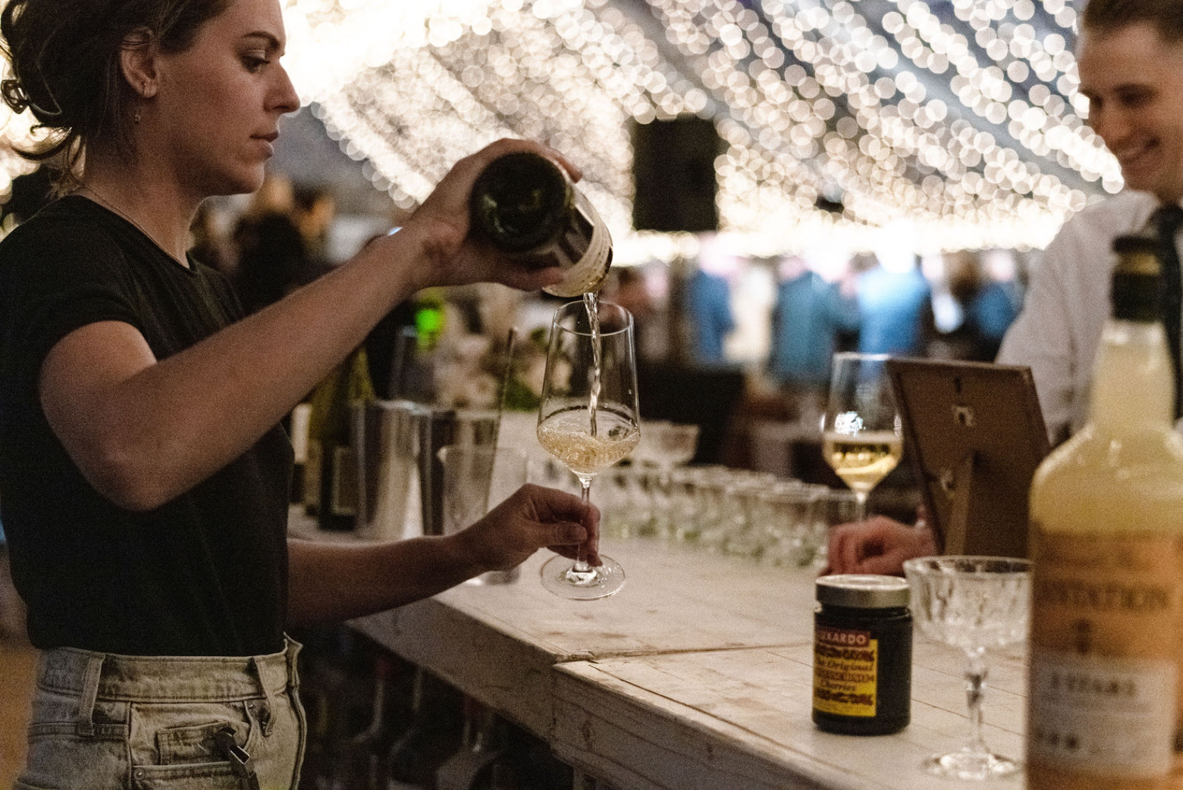A woman pours white wine into a glass at a bar, with a man smiling in the background. String lights decorate the ceiling.