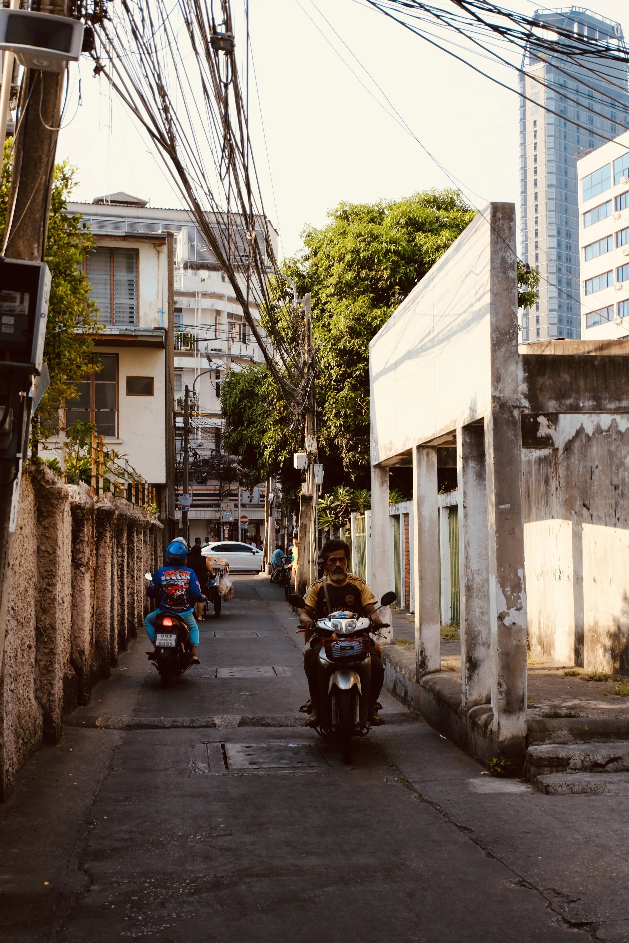 Street scene with two motorcyclists riding on a narrow road in an urban area, surrounded by buildings, trees, and power lines, with a clear sky.