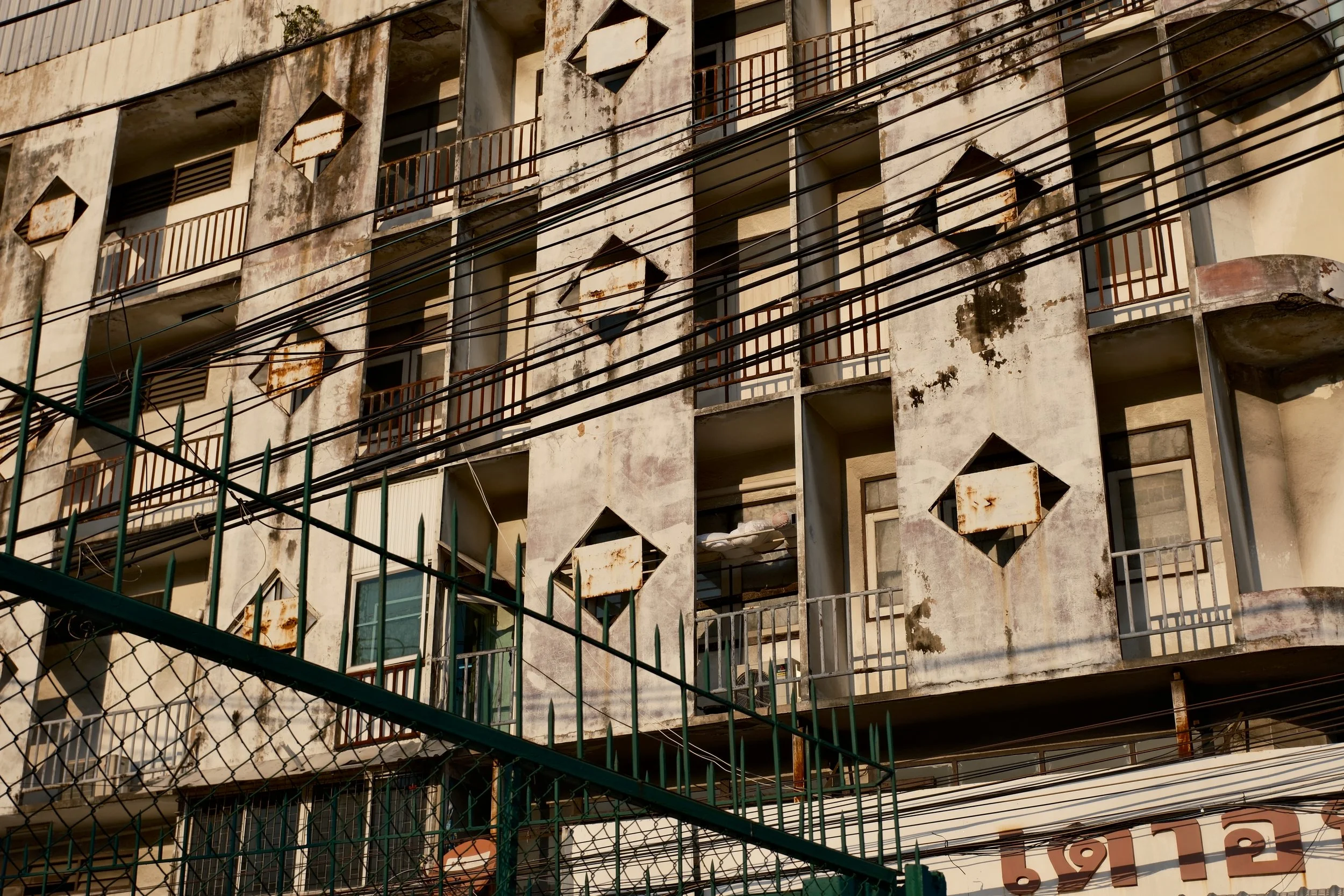 An old, weathered multi-story apartment building with rusted railings and peeling paint. The facade shows signs of neglect, with visible stains and damage. In front, there is a green metal fence and electrical wires crossing the scene.