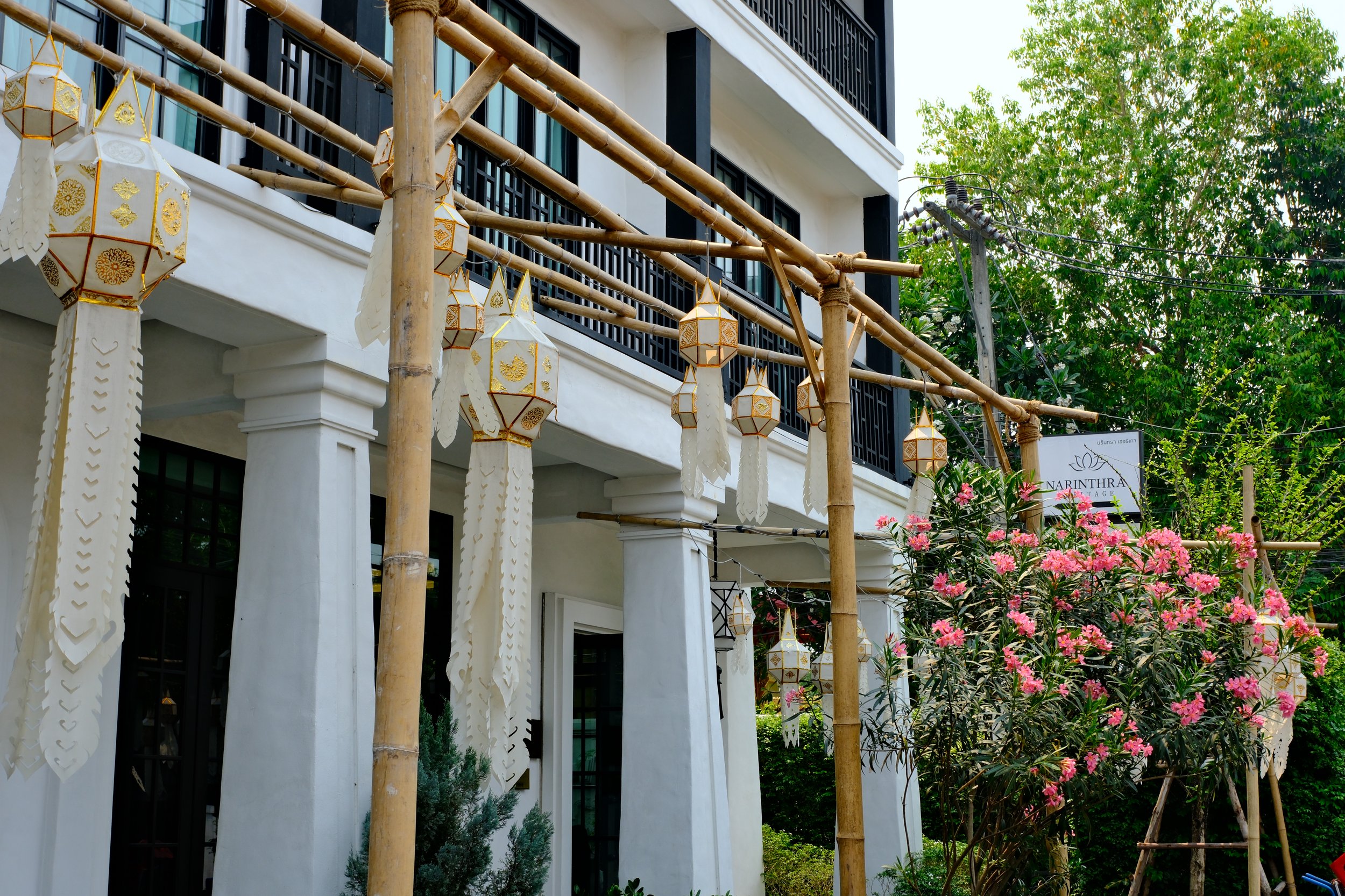 Exterior of a white building decorated with hanging paper lanterns and a flowering bush with pink blooms, bamboo poles supporting the decorations, and a sign that reads "Narinthra."