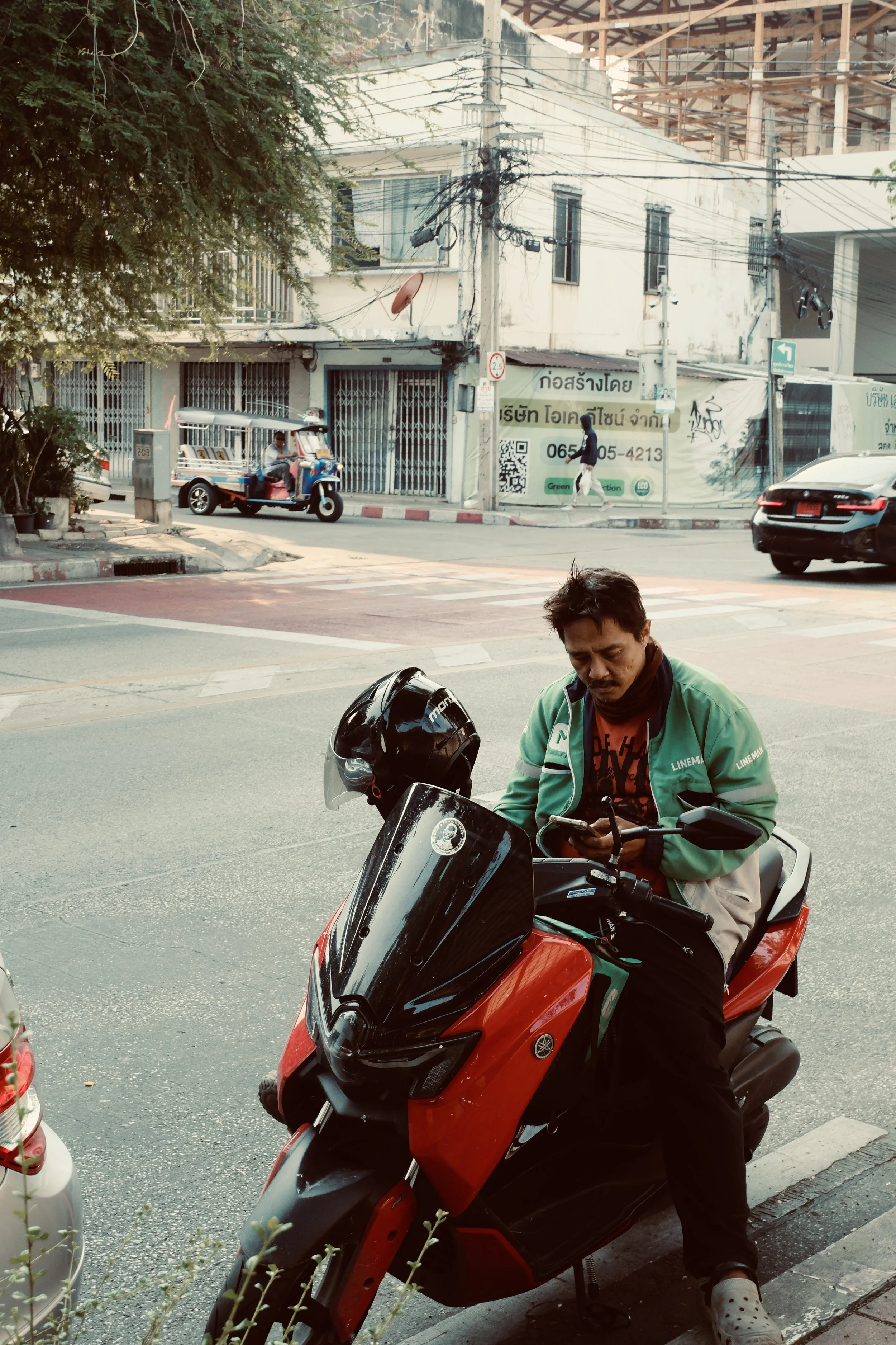 A man sitting on a red and black motorcycle, looking at his phone, on a city street with vehicles and pedestrians in the background.