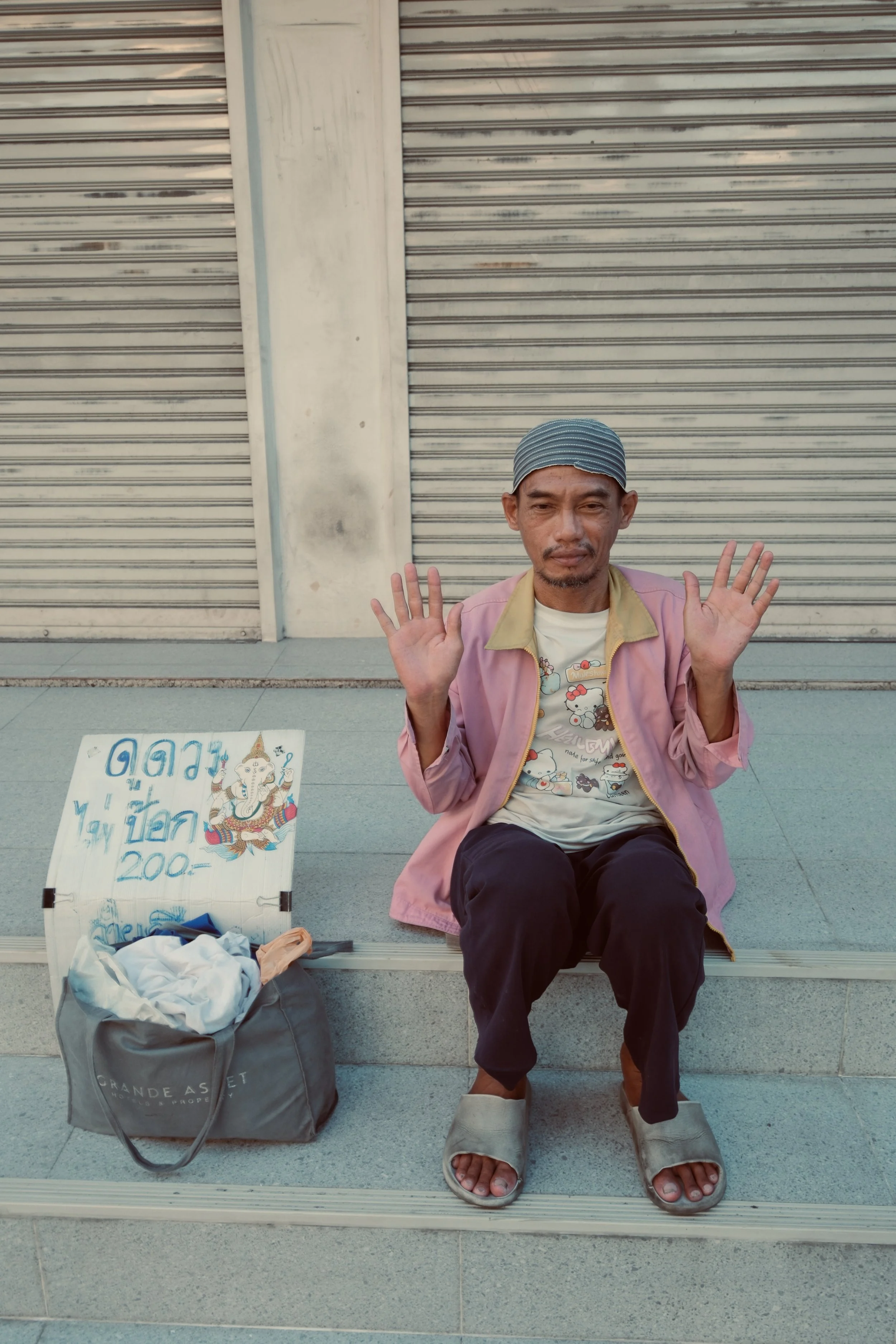A man sitting on a step with his hands raised. Next to him is a bag and a sign written in Thai.