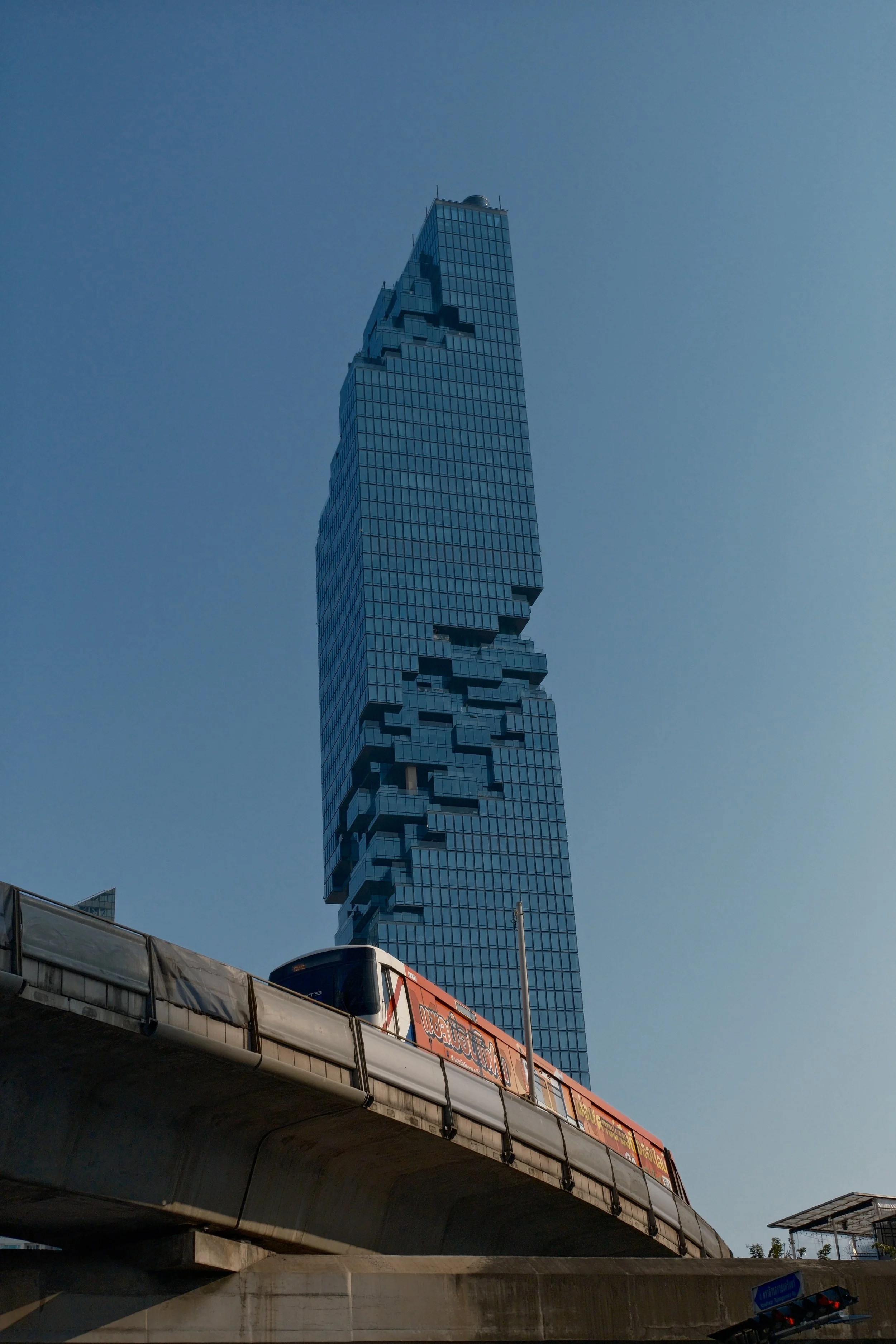 A tall modern glass skyscraper with a unique, pixelated design partially obscured by an elevated train track and a moving train in the foreground.