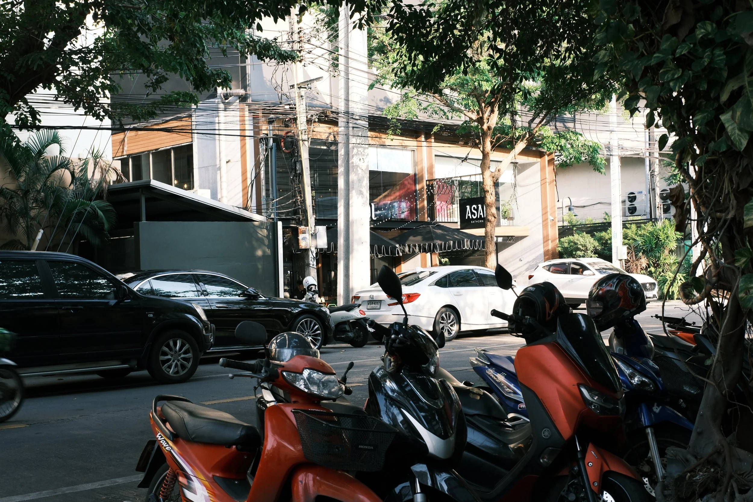 Street view showing parked cars and motorbikes with a modern building in the background, trees, and power lines.