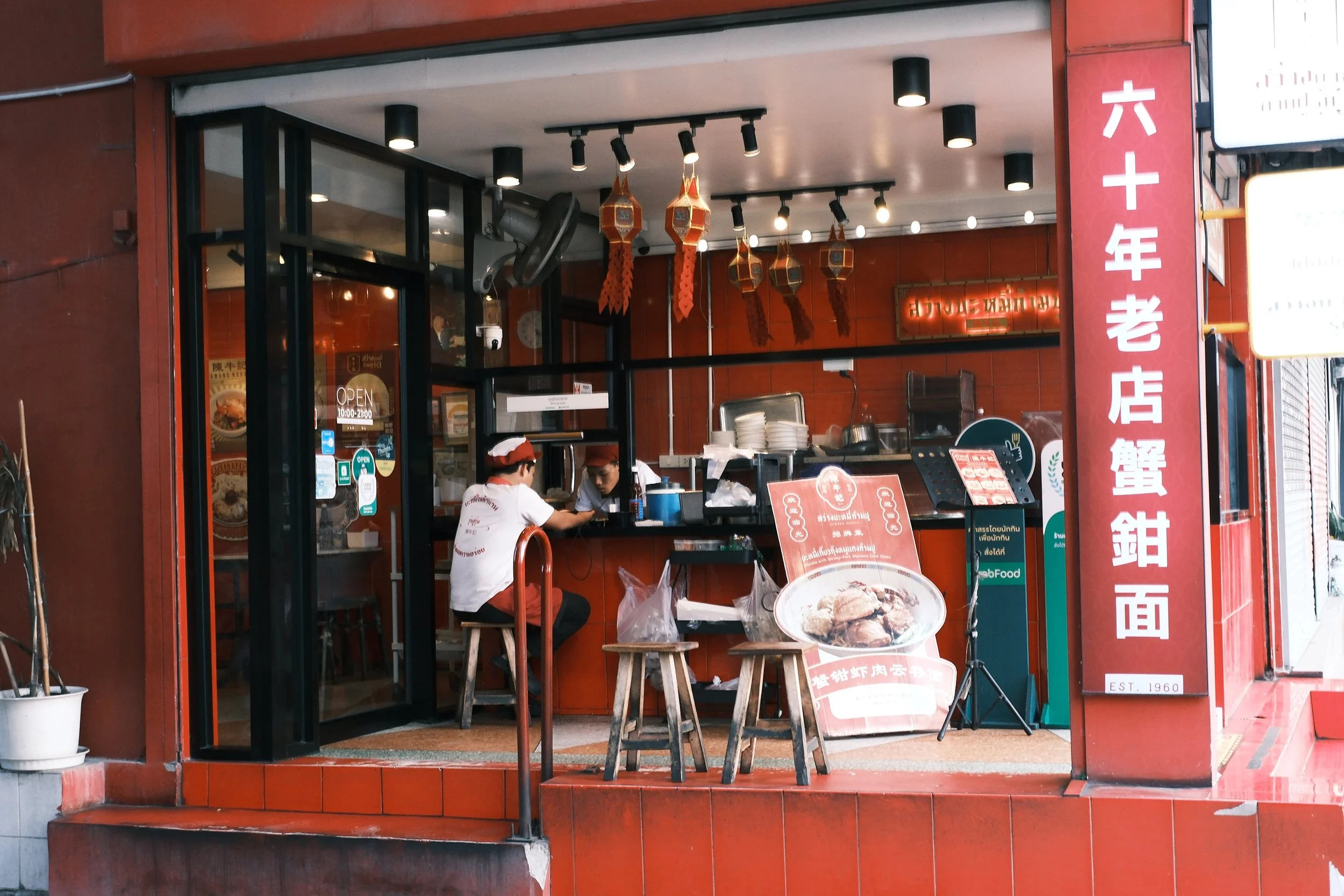 Small Asian restaurant with red exterior and Chinese characters on a sign. Inside, two staff members wearing hats work behind the counter. Hanging lanterns decorate the ceiling, and a menu board features a picture of a dish. The restaurant has an ope