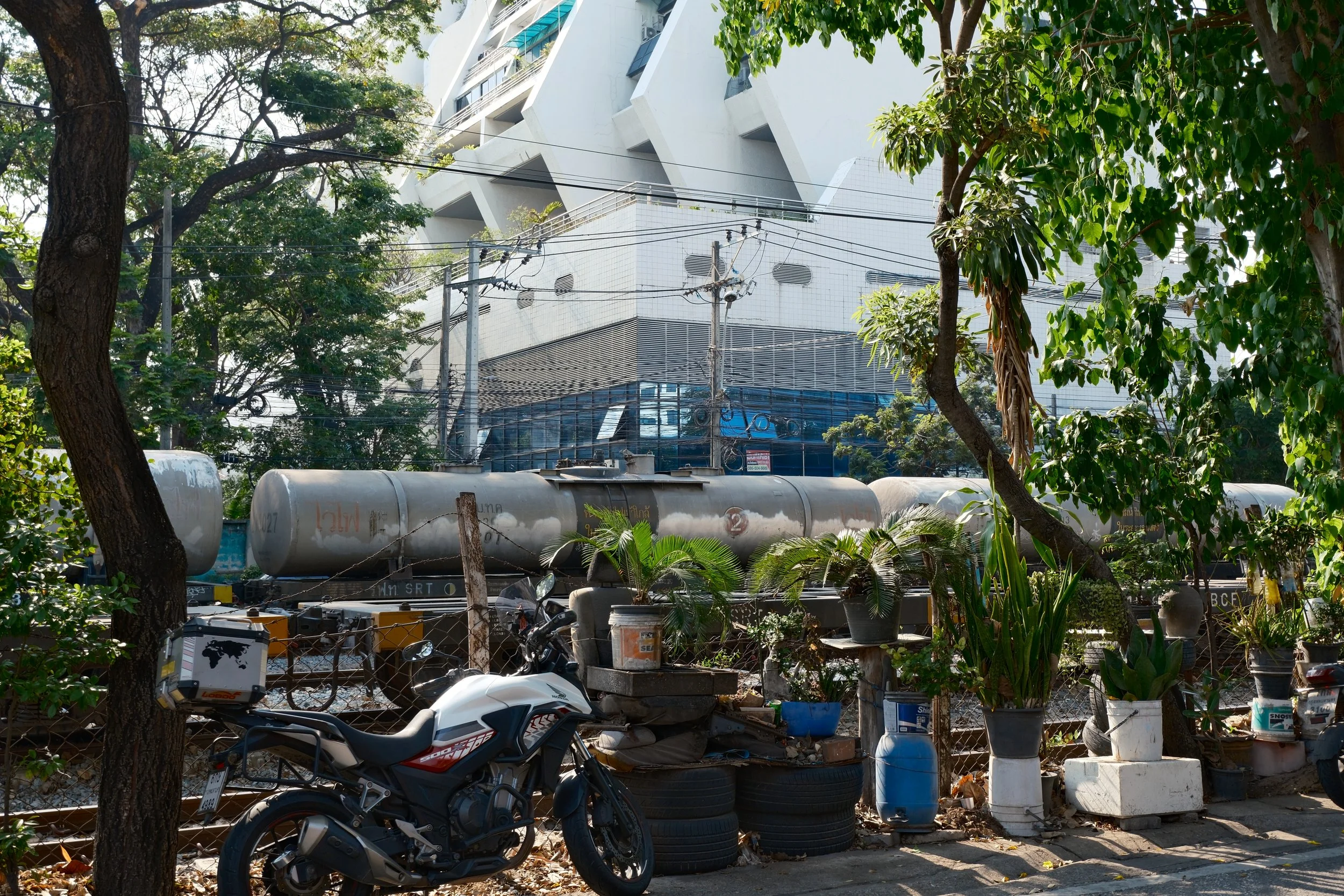 An urban scene with a motorcycle in the foreground, plants in pots and tires, a fence, graffiti, and a large modern building in the background.