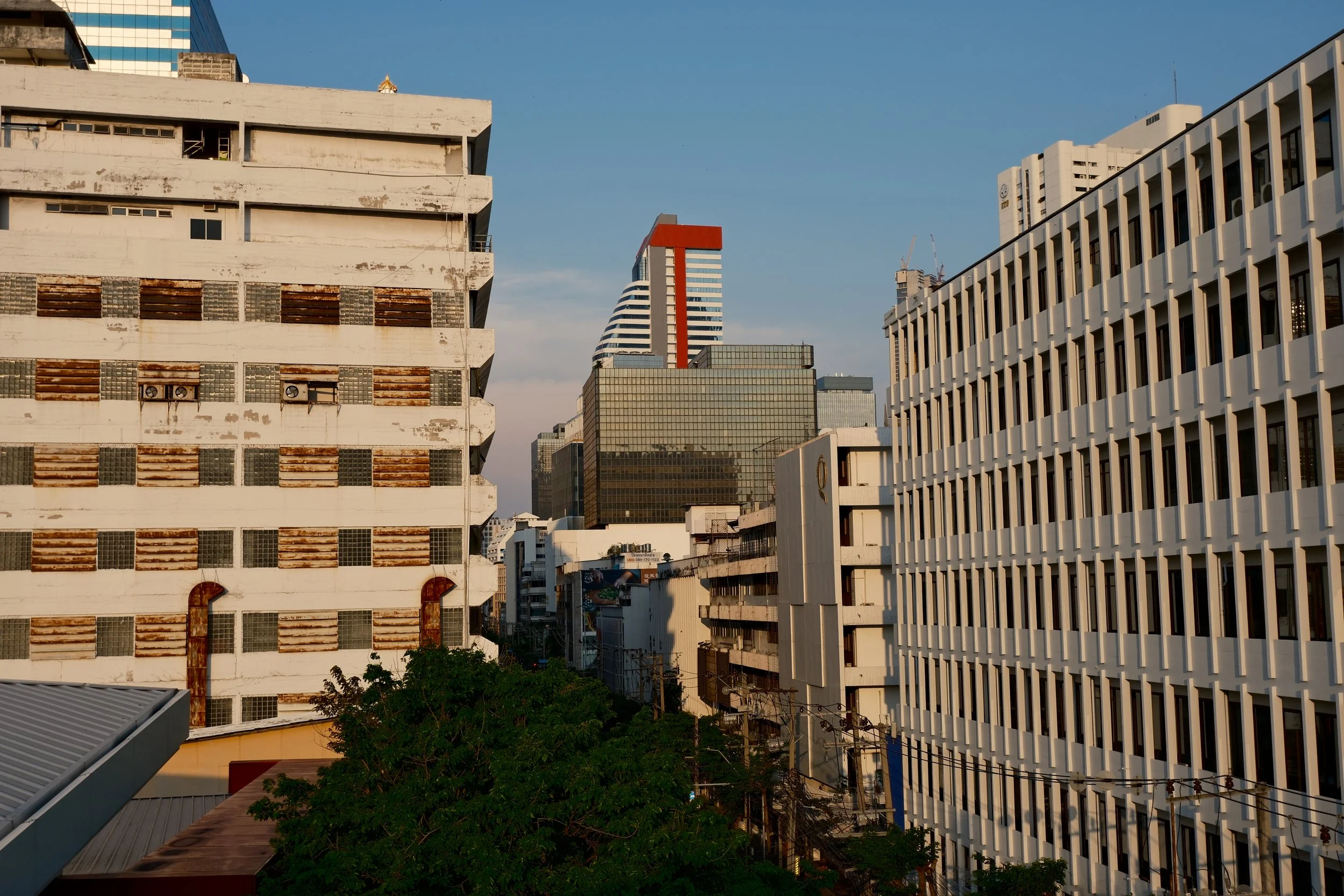 A cityscape featuring old and modern buildings with a blue sky in the background. The image includes a weathered white building with rusty pipes on the left, a black glass skyscraper with a red structure on the top center, and a white building with multiple windows on the right. Green trees are visible at the bottom.