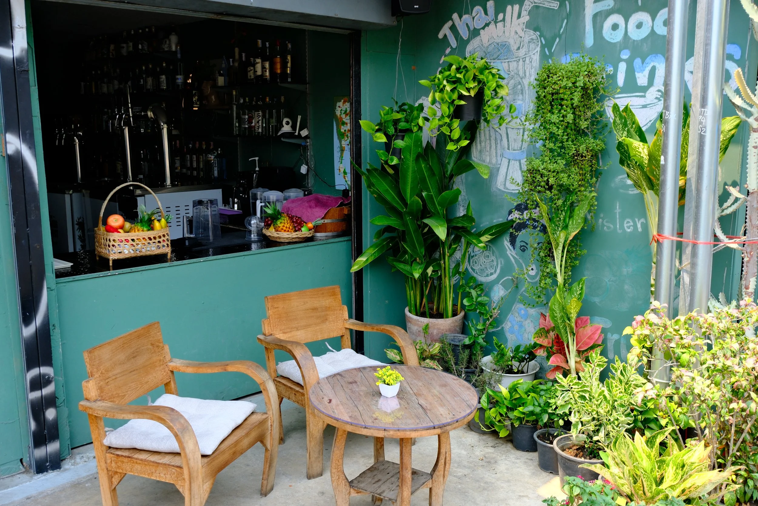 Outdoor seating area with two wooden chairs, a small round table holding a potted plant, surrounded by various potted plants and greenery, with a food stall or bar in the background and chalkboard art on the wall.